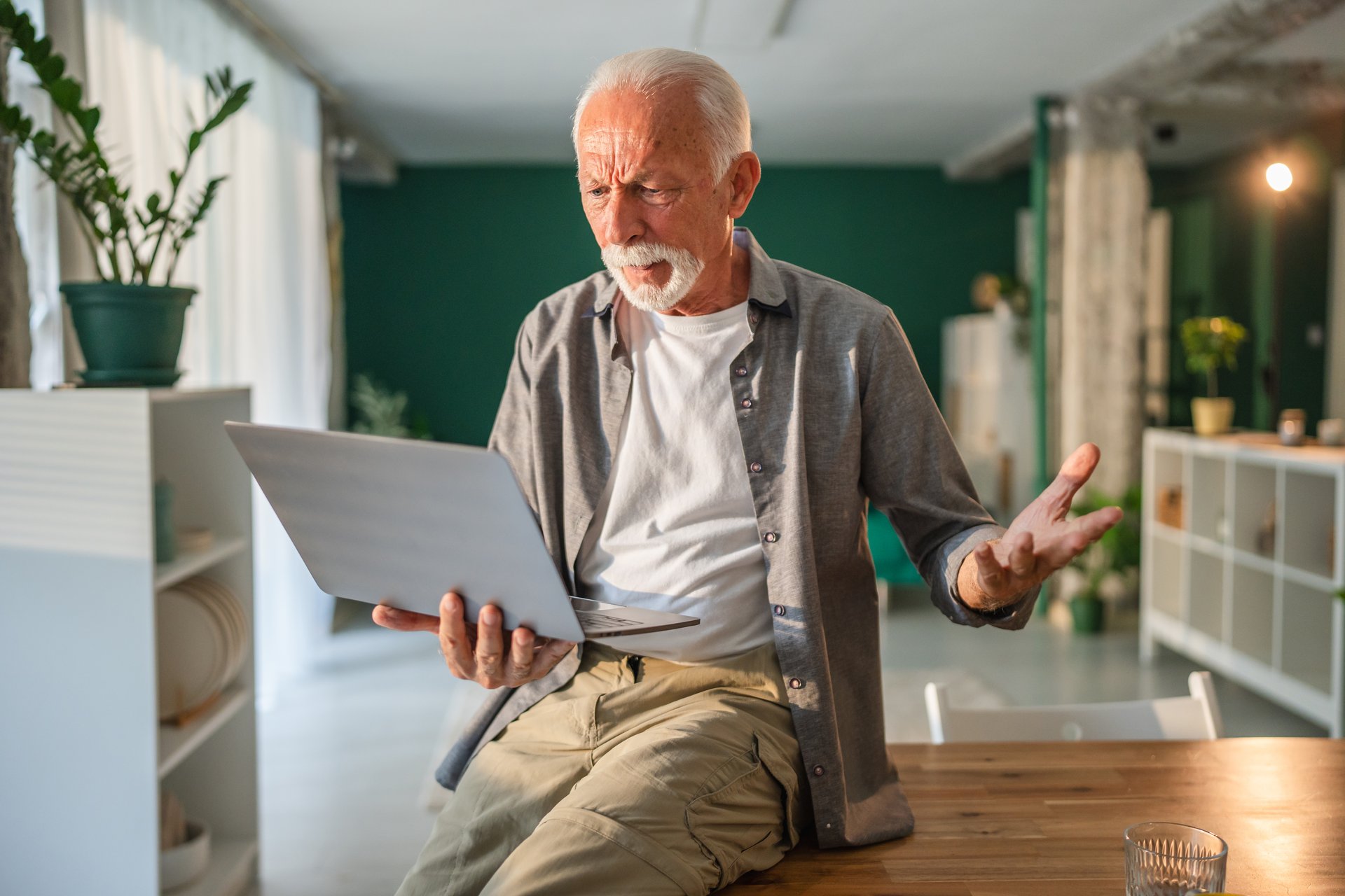Senior man getting frustrated while working from home, holding a laptop and gesturing with his hand, showing his confusion and annoyance with technology