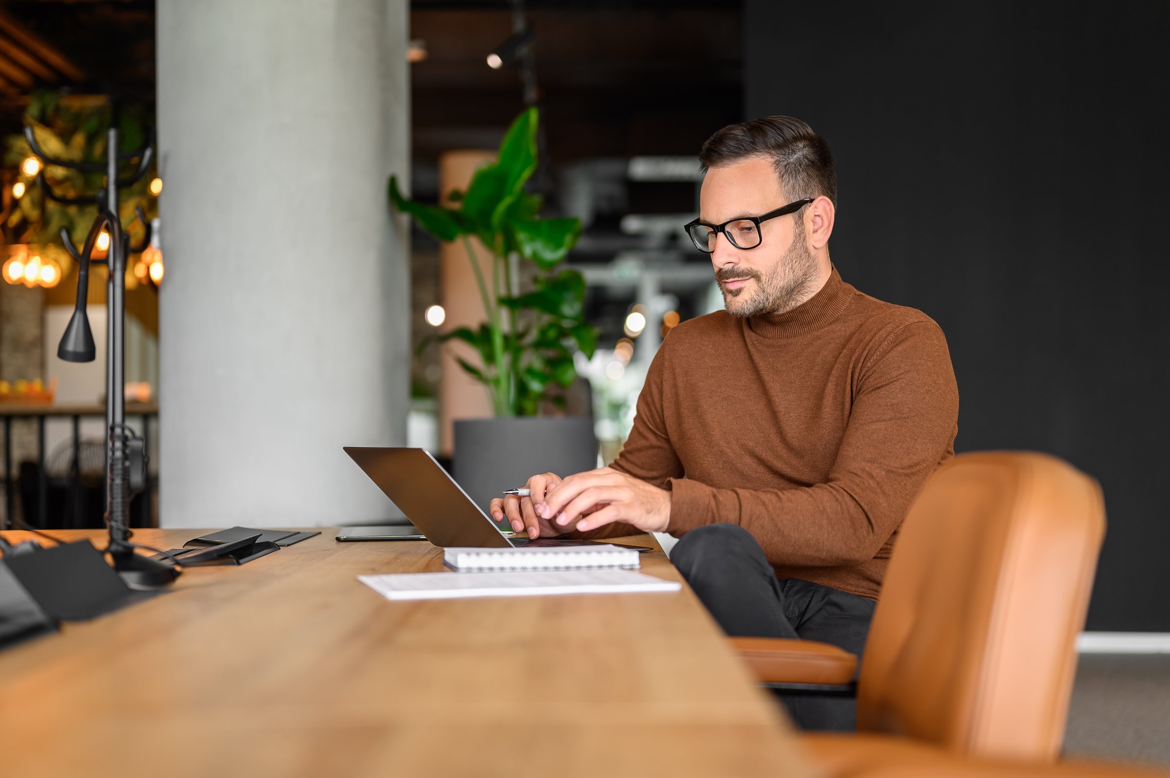 Focused male programmer in eyeglasses working serious on project over computer at desk in office