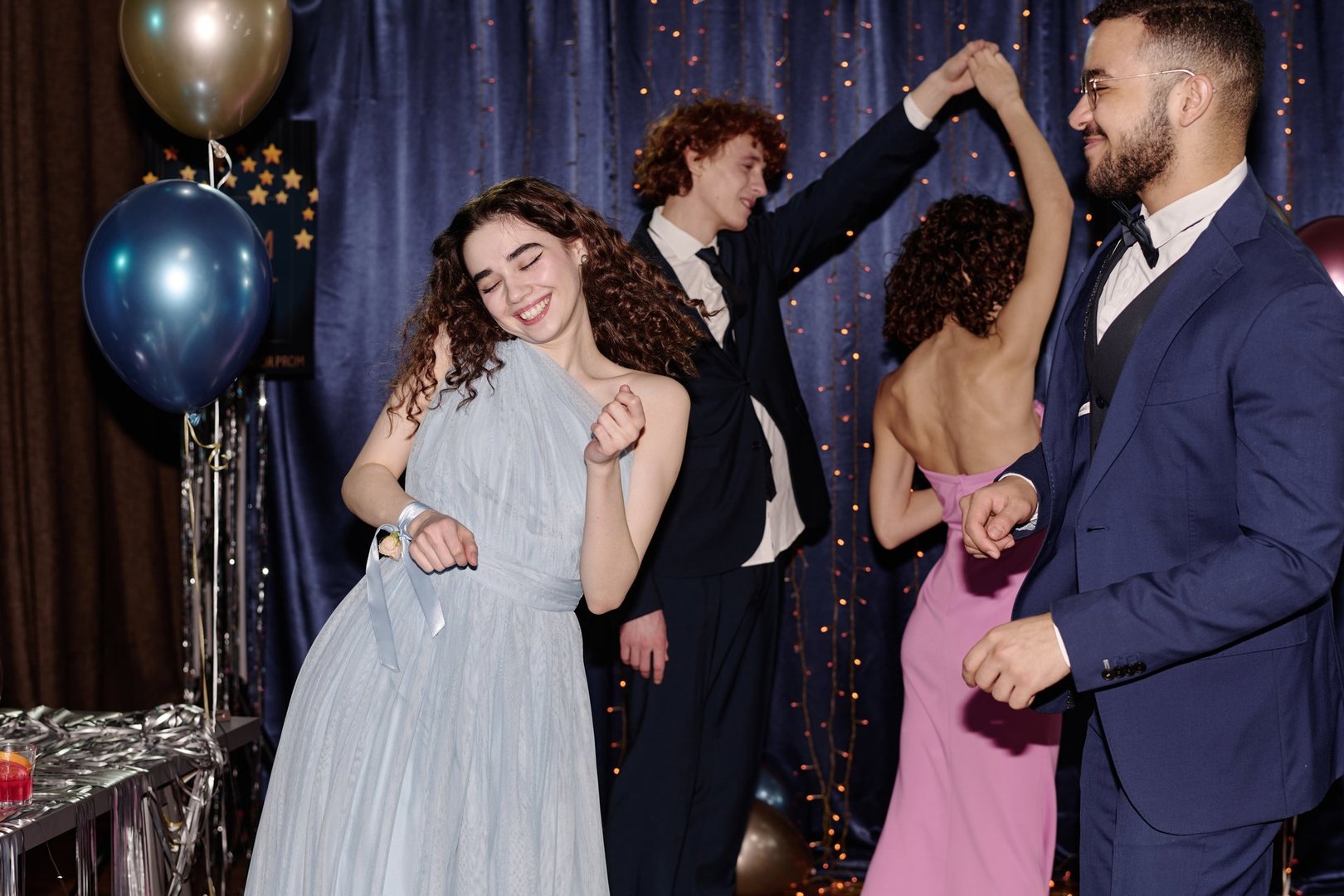 Caucasian teenage girl dancing joyfully at party with friend smiling nearby, teenagers in formal attire dancing together in background, festive decorations visible