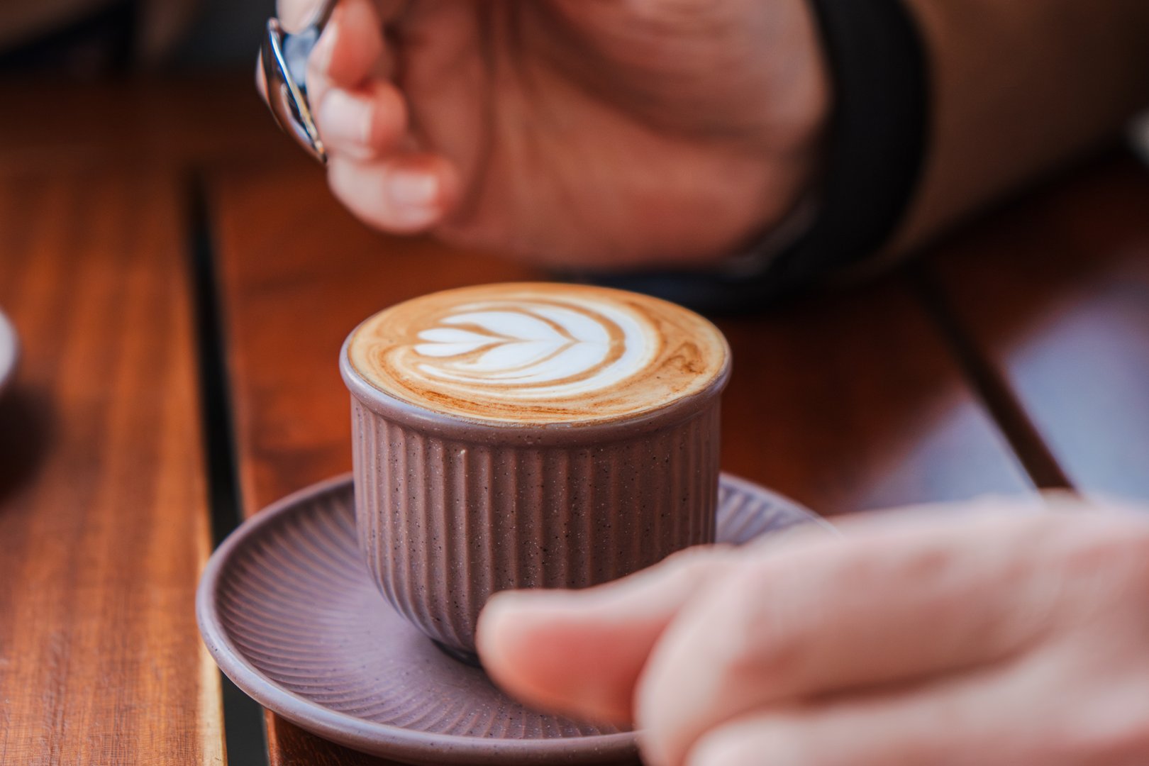 A beautifully crafted latte art displayed in an elegant cup, with a hand holding a spoon ready to stir. The intricate leaf patterns on the creamy surface highlight the artistry of coffee making and enjoyment.