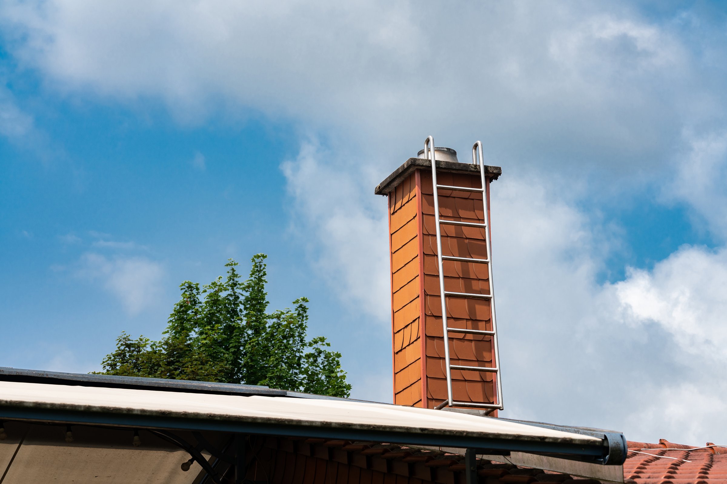 A tall red brick chimney rises against a backdrop of bright blue sky and fluffy clouds. Lush green trees surround the structure, indicating a residential environment.
