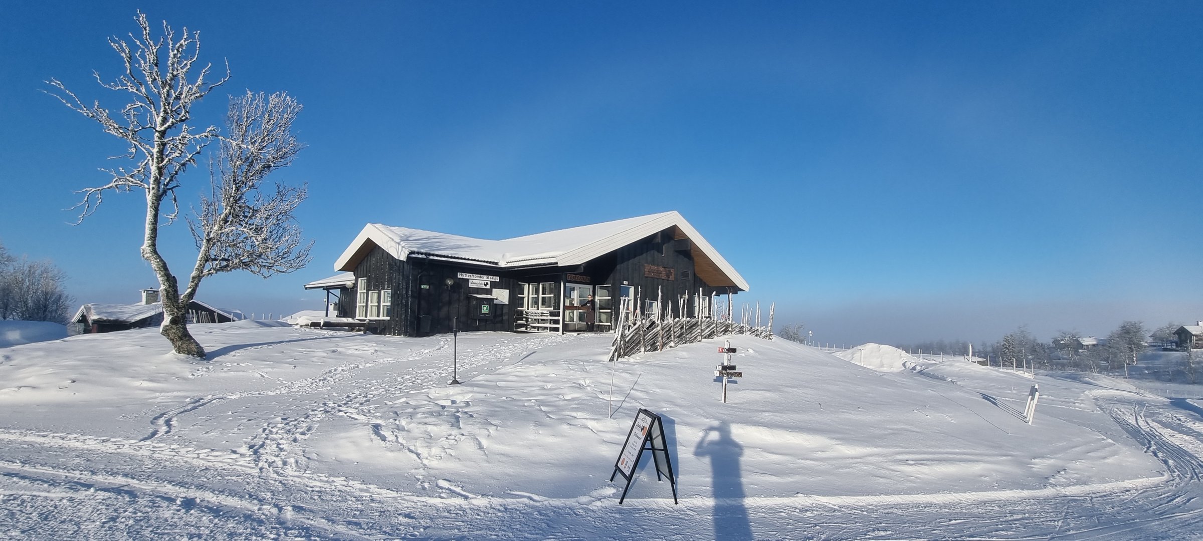 A snowy landscape with a wooden cabin, signage, and a lone tree on a clear day; a shadow stretches across the snow.