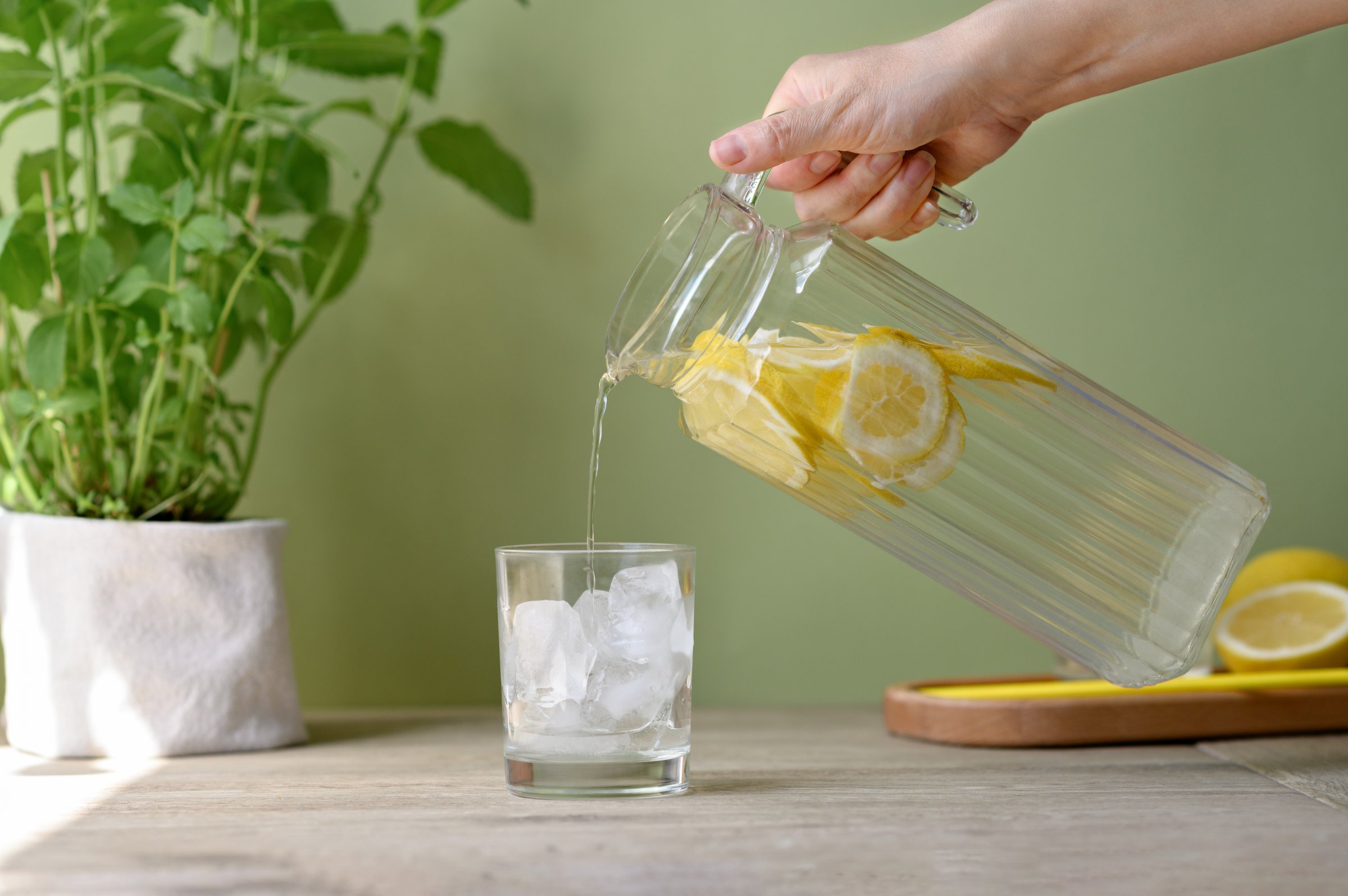 Hand holding jug with lemonade drink pouring beverage in glass with ice, soft green background, plant and lemons on kitchen table, summer non alcohol healthy drink.