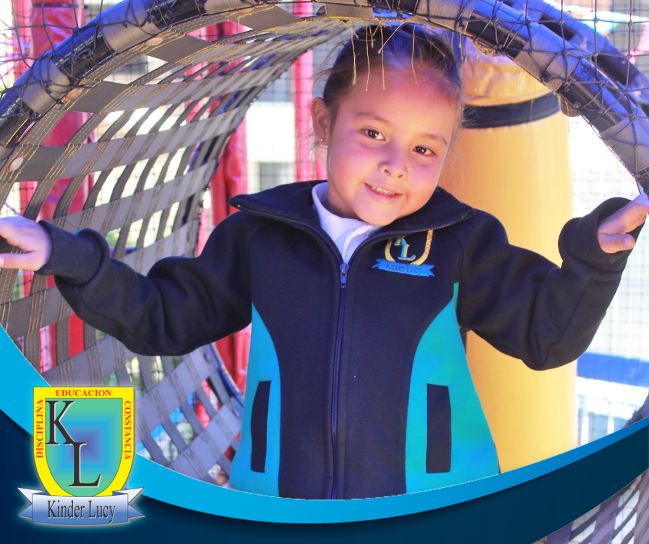 Young child smiling inside a play area with a netted tunnel, wearing a dark jacket with a Kinder Lucy logo.
