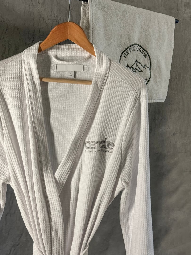White bathrobe on a hanger with "Cenote" logo, hanging in front of a towel with "Arctic Oasis" emblem.