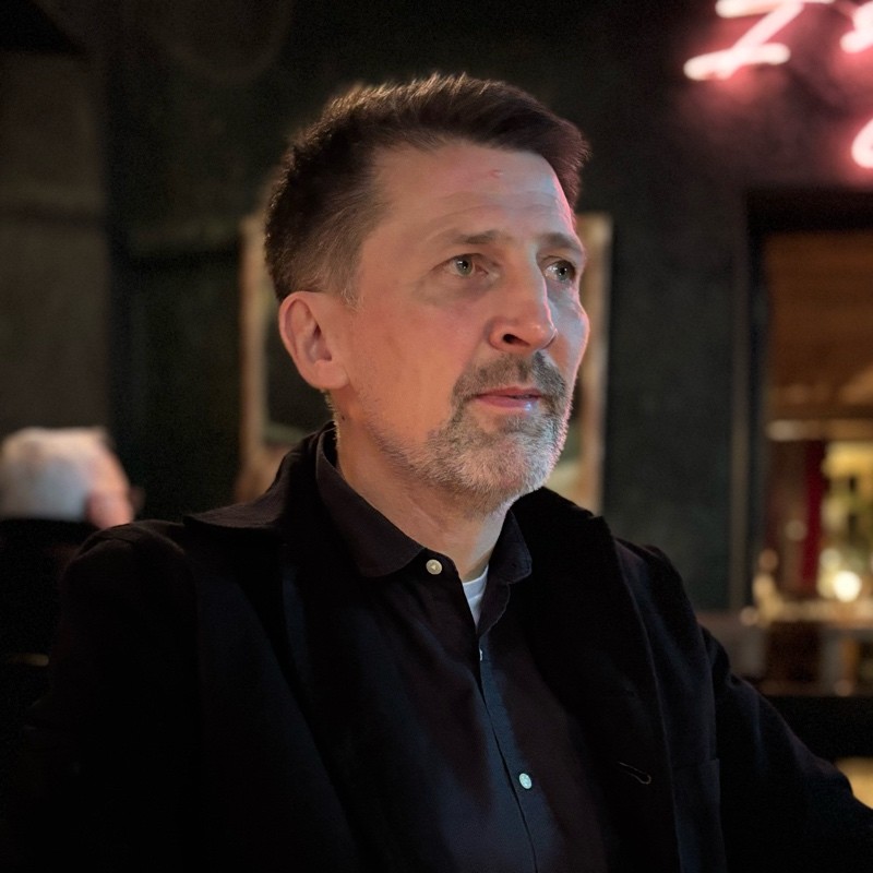 Man with short dark hair and beard in a dimly lit room, wearing a black jacket and shirt, looking thoughtfully to the side.