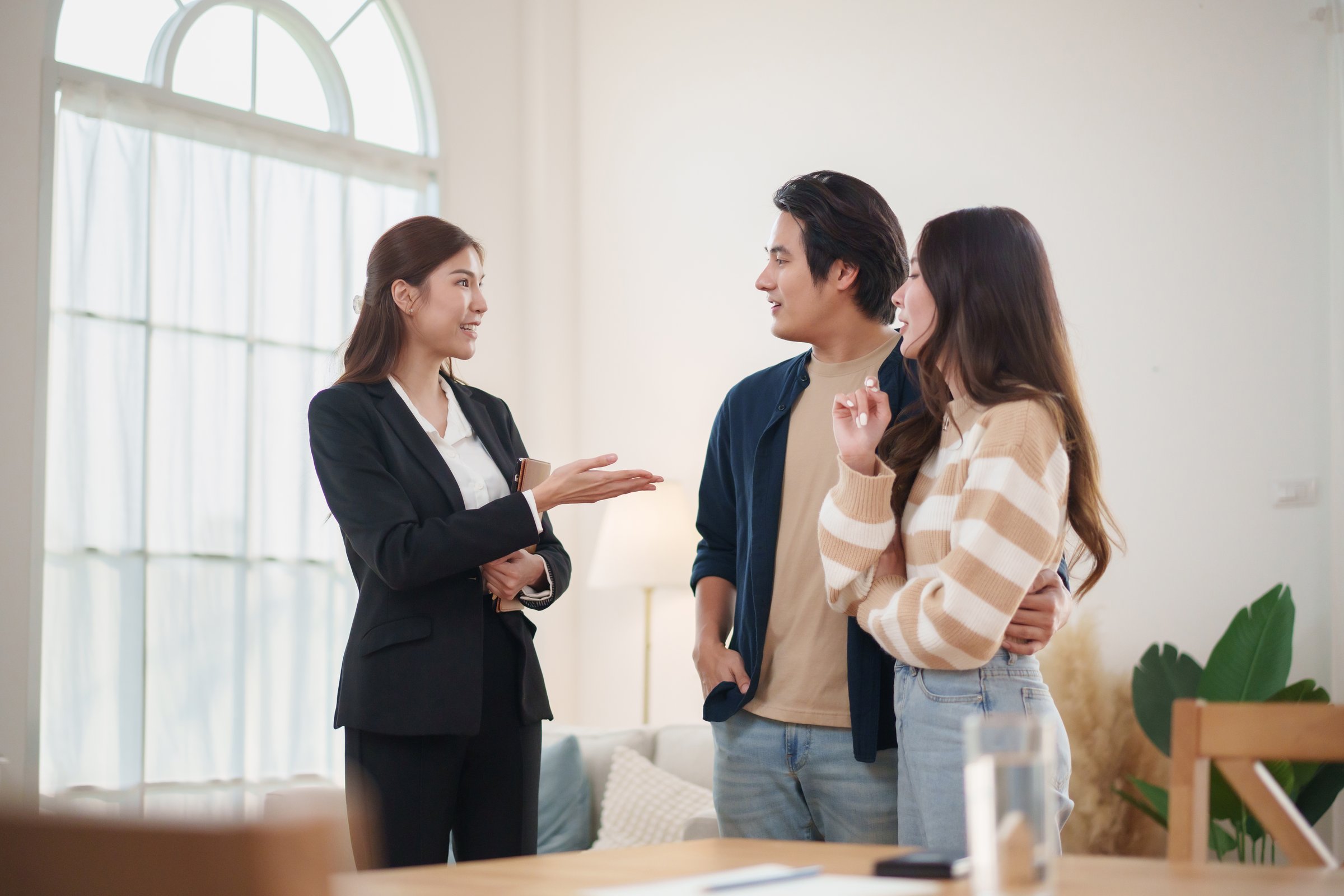 Asian Female real estate agent explaining house details to happy couple in casual outfits standing together.