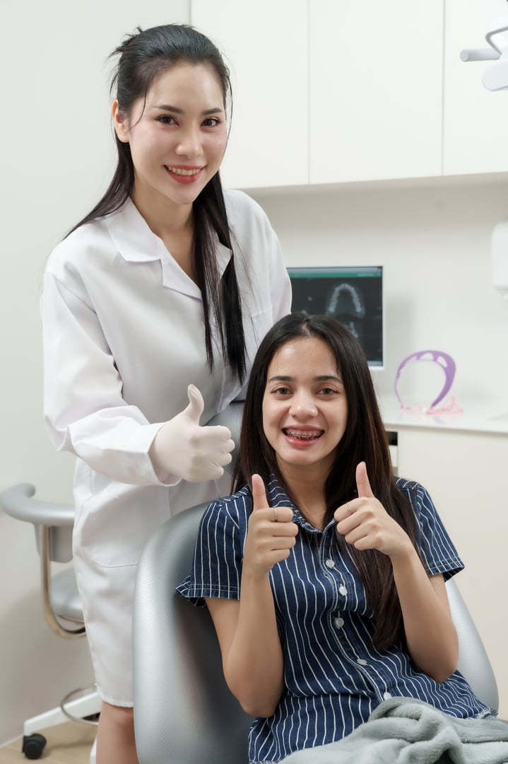 Asian female dentist adult and asian female patient adult sitting on dental chair smiling confidently and giving double thumbs up after dental treatment in clinic with x-ray screen and tools