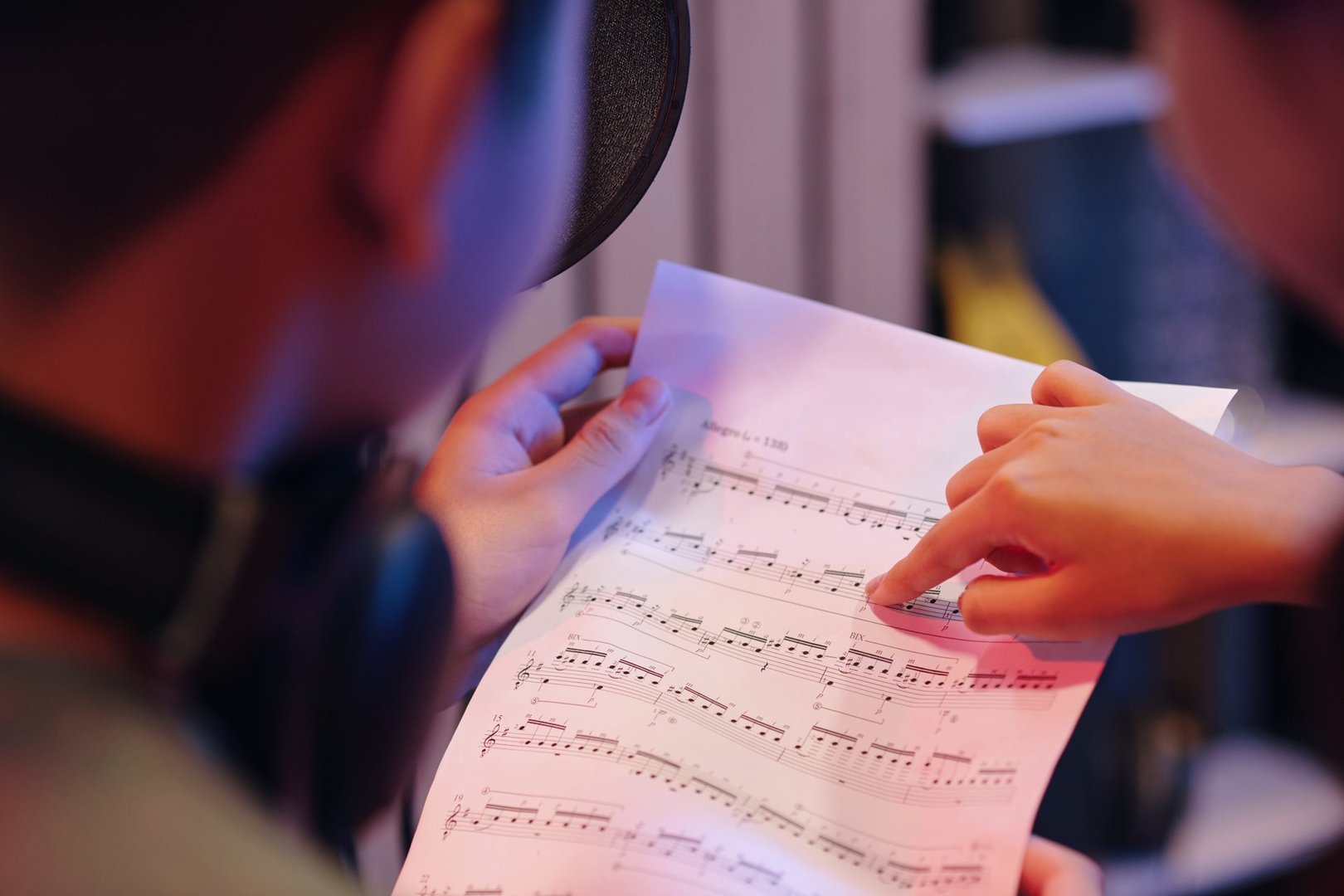 Close-up of two people reviewing sheet music with one pointing at notes in professional recording studio. Microphone and other equipment visible in background