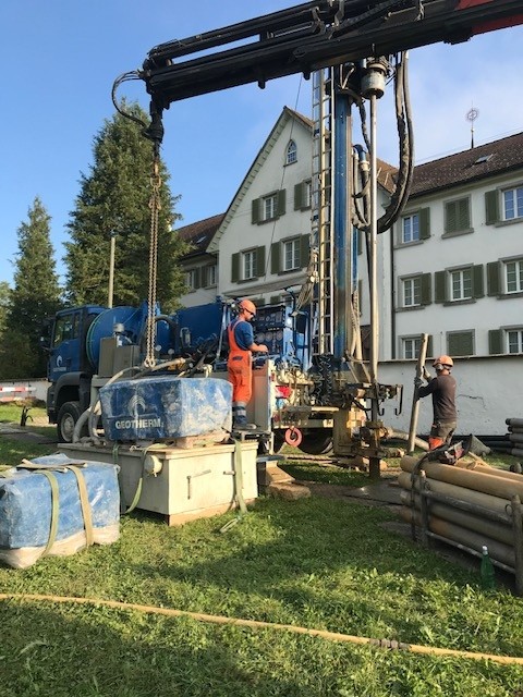 Two workers operate a large drilling machine near a building, surrounded by equipment and greenery on a sunny day.