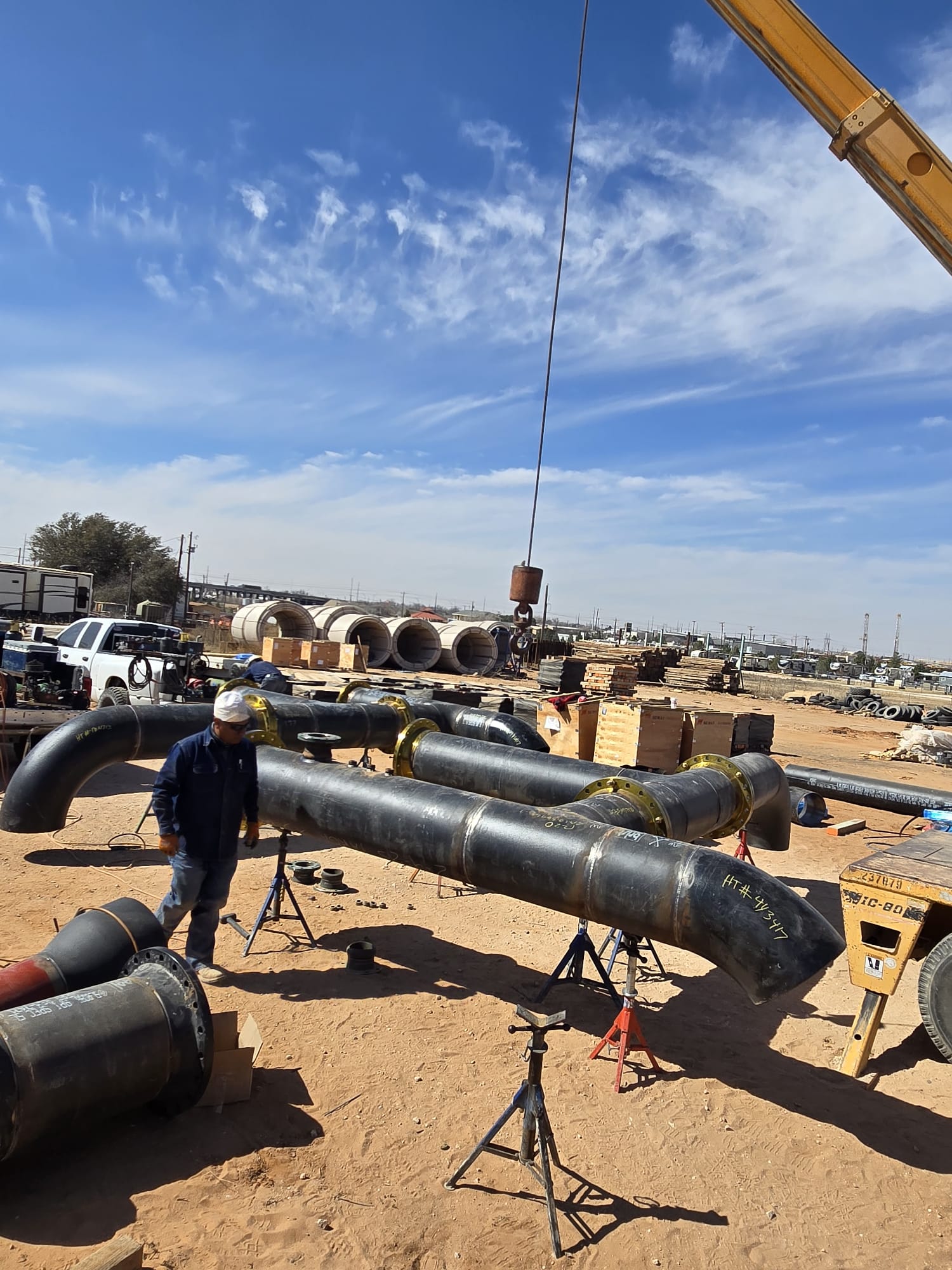 Construction site with large pipes on stands, a worker in safety gear, and a crane lifting equipment on a clear day.