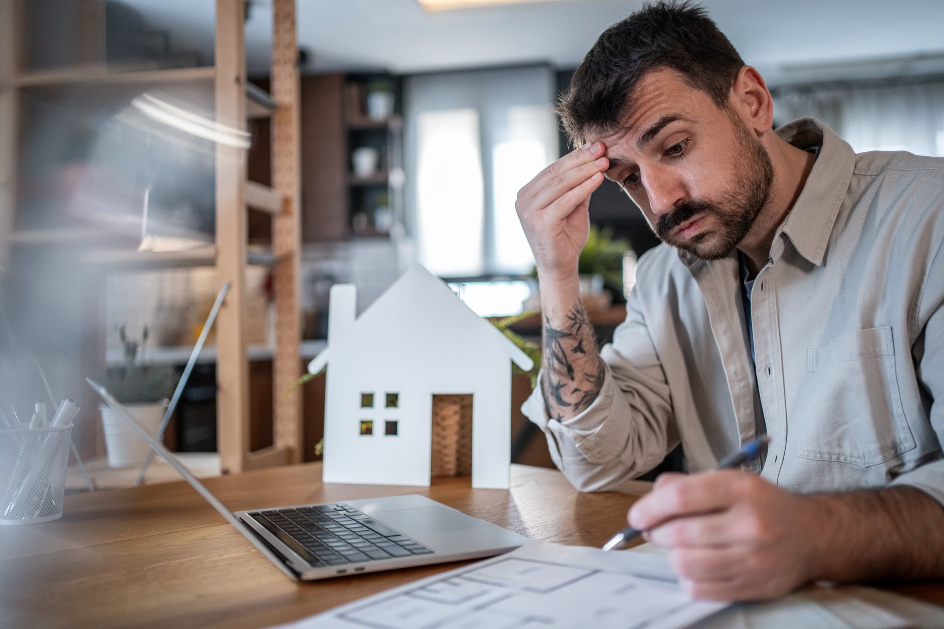 Architect working from home, reviewing blueprints and using a laptop, feeling overwhelmed by stress and fatigue from the demands of design and project responsibilities