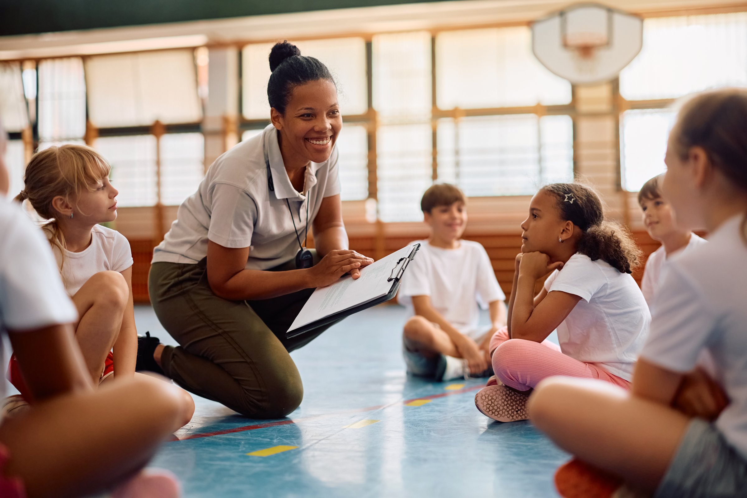 Happy African American sports teacher and her elementary students talking at school gym.