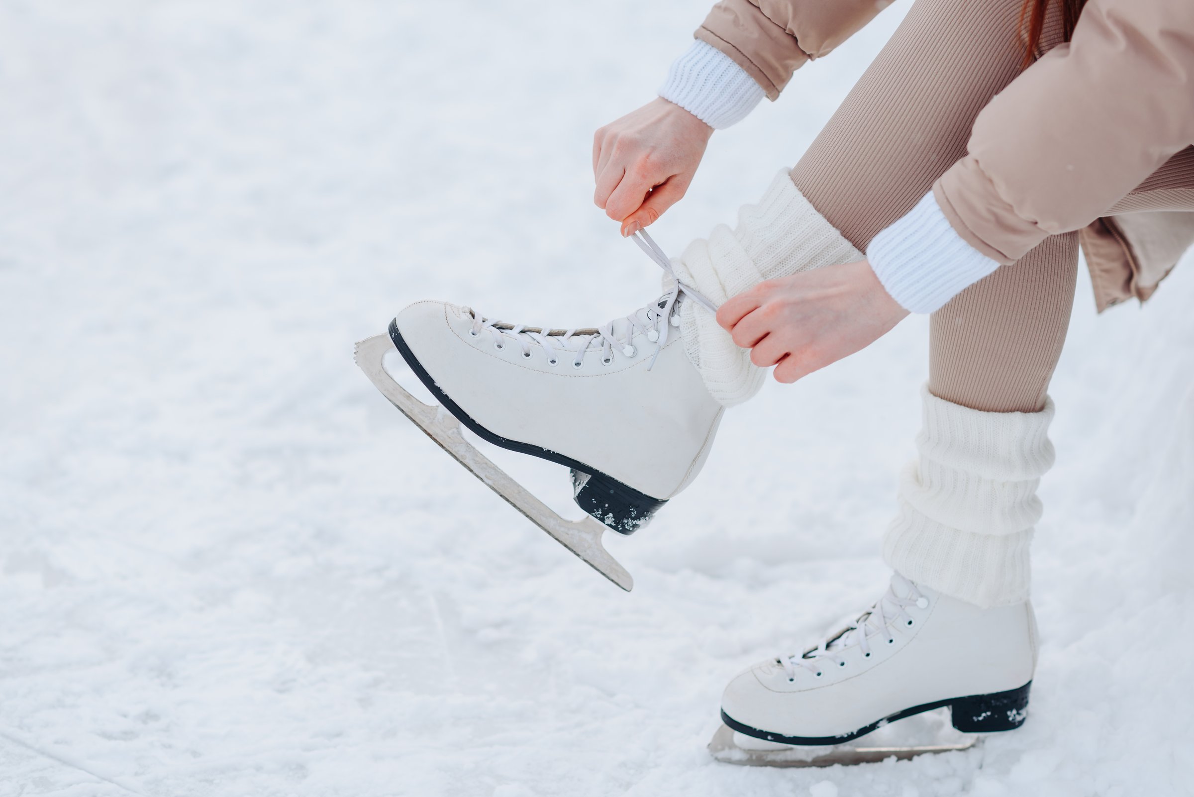 young girl in white and beige winter clothes lacing up skates before skating in ice rink in winter cloudy snowy day, close-up view of skates, copy space