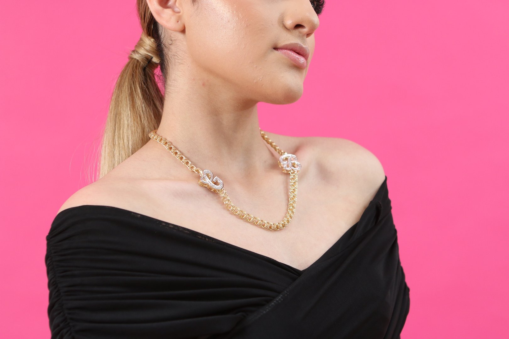 Woman wearing a gold chain necklace with shiny details, off-the-shoulder black top, and pink background.
