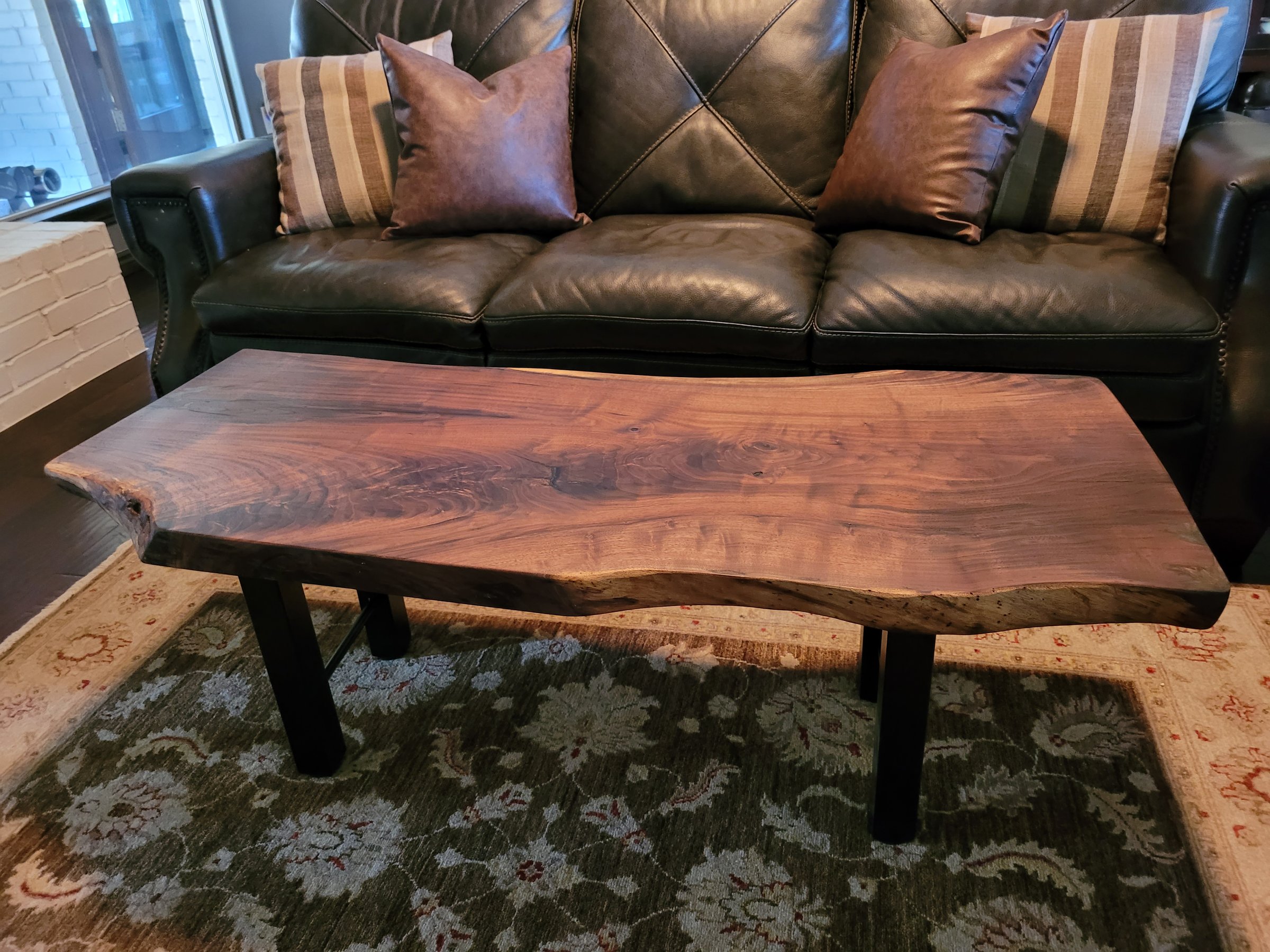 A wooden coffee table with a natural, live edge design, positioned in front of a black leather sofa with decorative pillows.