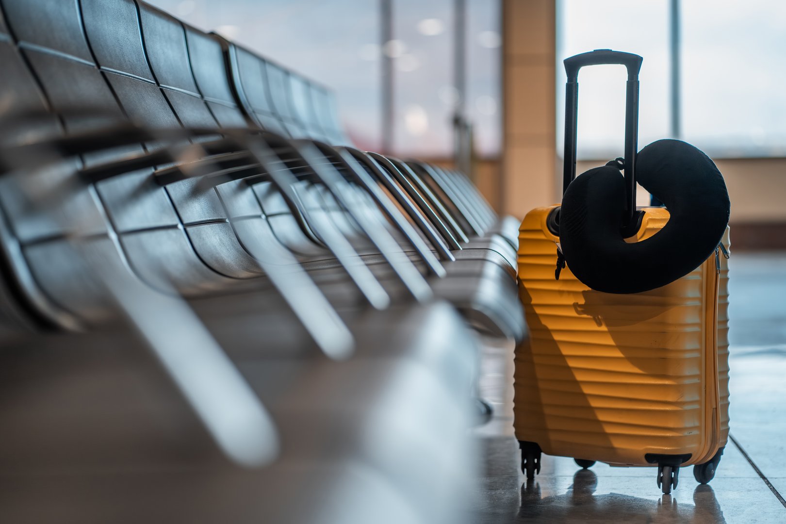 portable and stylish yellow suitcase with neck pillow rests beside empty airport seatings row as travelers wait at busy terminal, baggage at travel and vacations concept. High quality photo