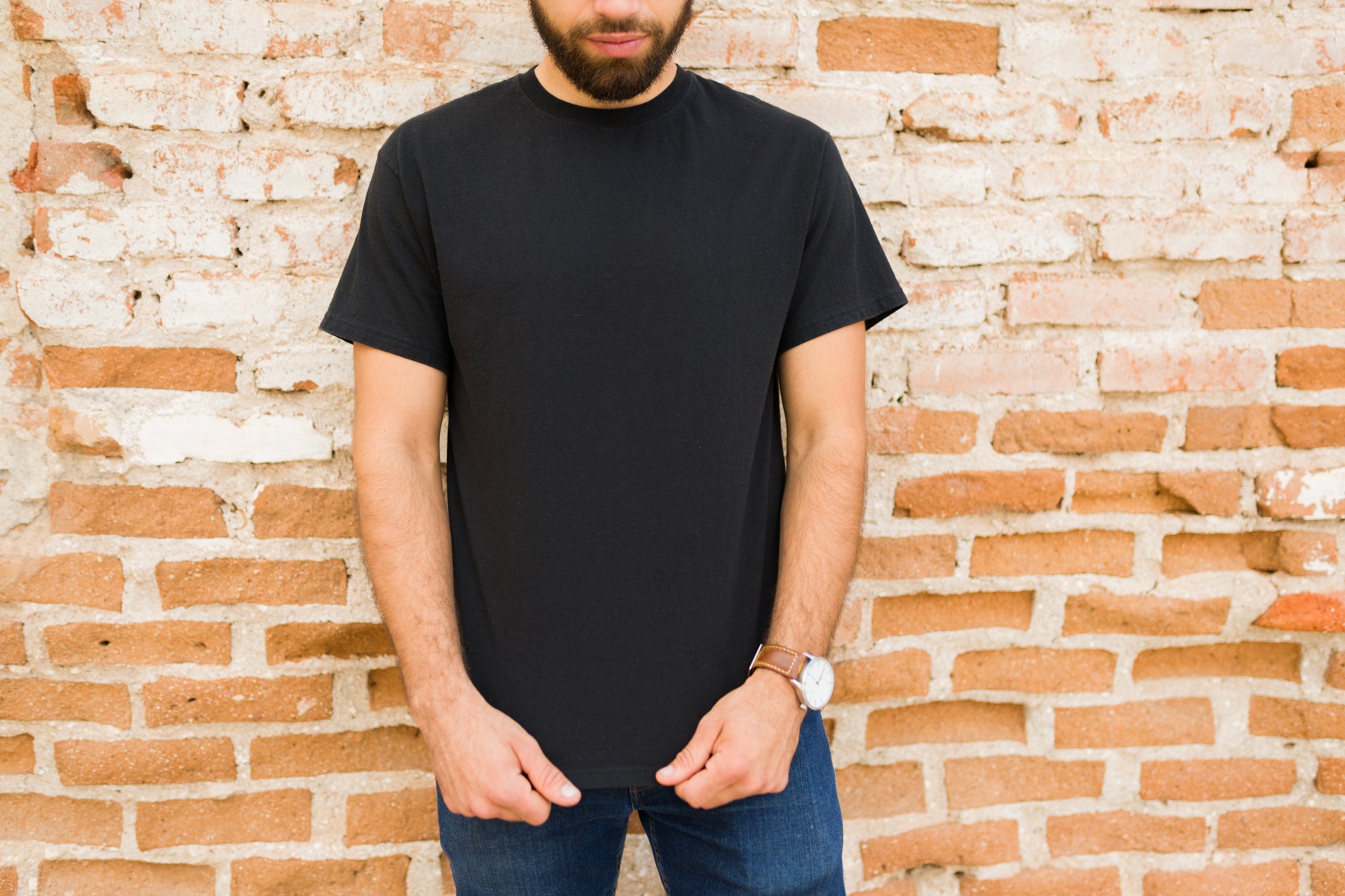 Attractive man wearing a blank black t-shirt ready for branding mockup stands confidently in front of a rustic brick wall
