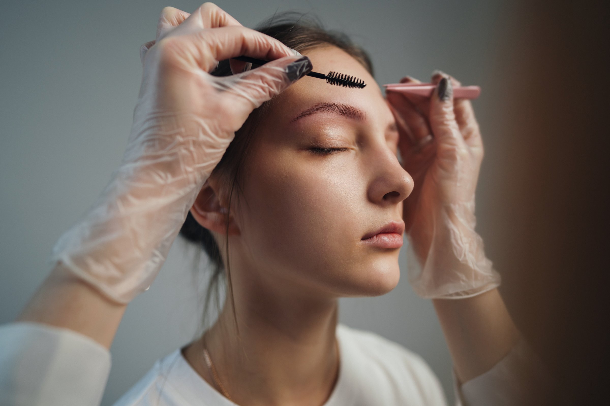 Gloved esthetician performing eyebrow shaping procedure with precision tools, sterile gloves, client eyes closed