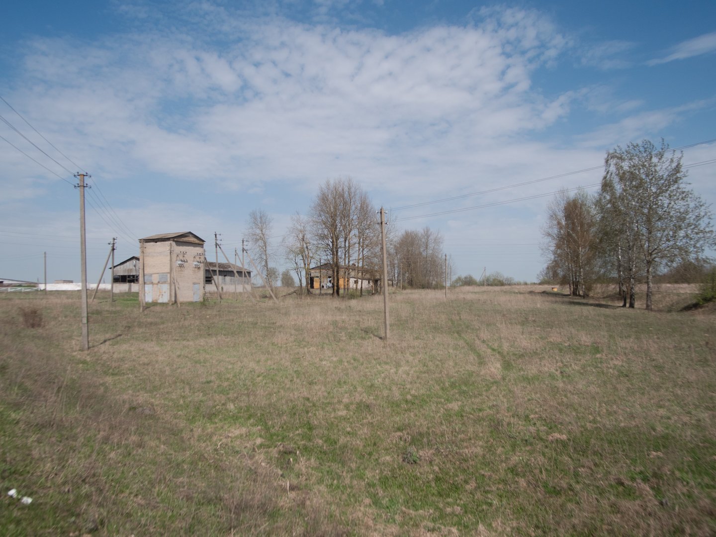 Several old, abandoned buildings stand isolated in a grassy field under a bright blue sky with scattered clouds, indicating the arrival of spring.
