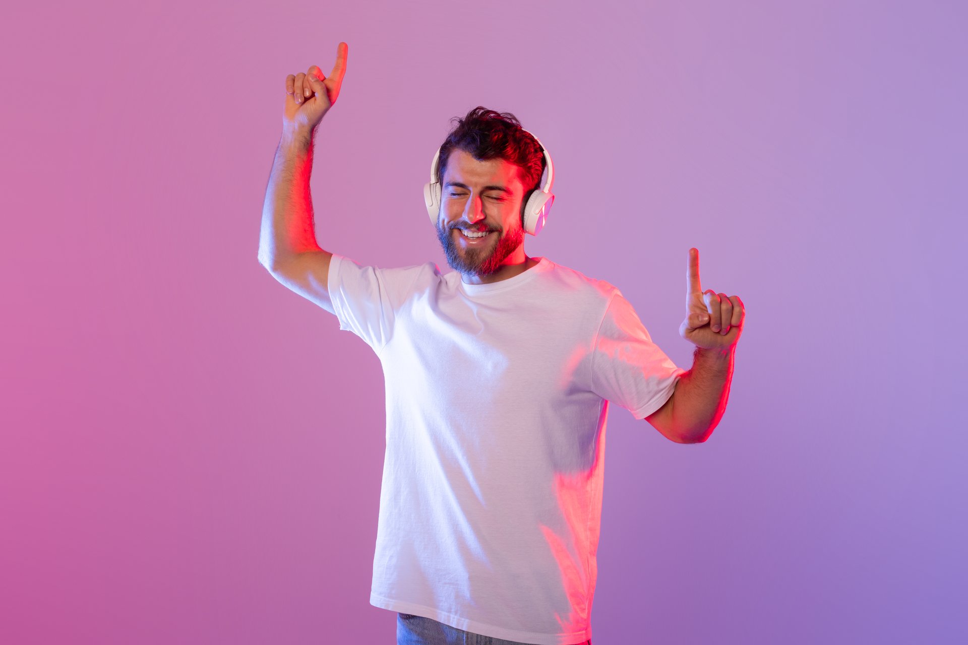 A cheerful man wearing wireless headphones is dancing and enjoying music. The background features vibrant, colorful studio lighting, enhancing the lively atmosphere.