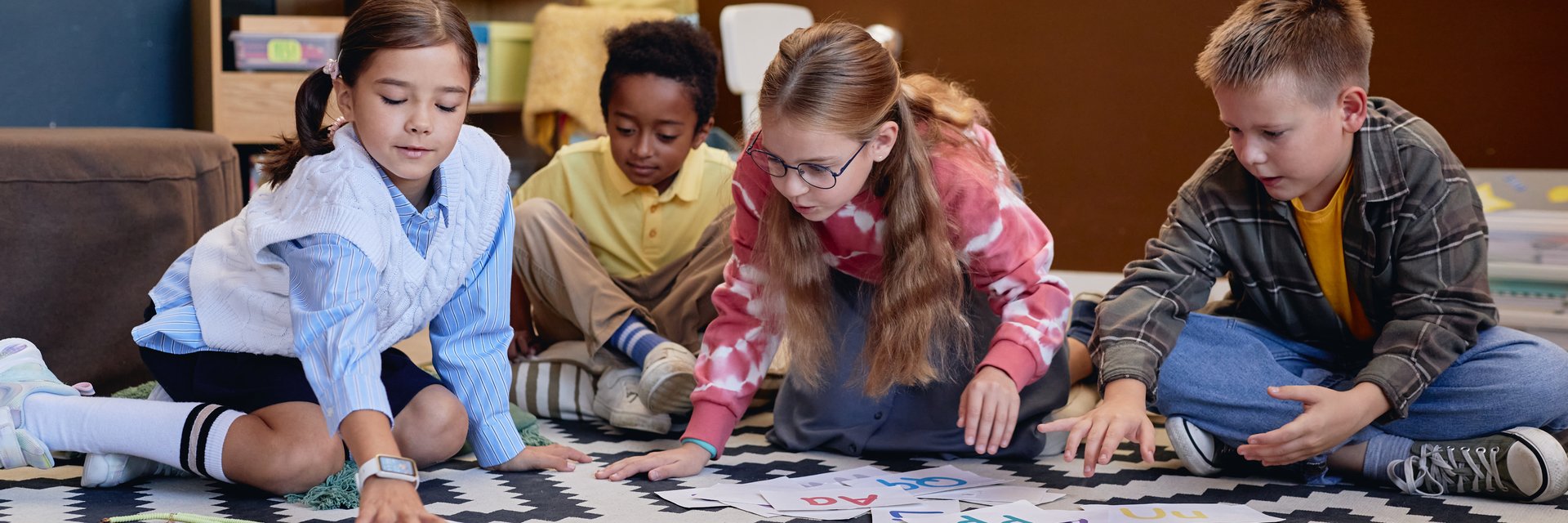 Header image of multiethnic group of children learning English alphabet and playing with letter cards sitting on floor in school classroom together