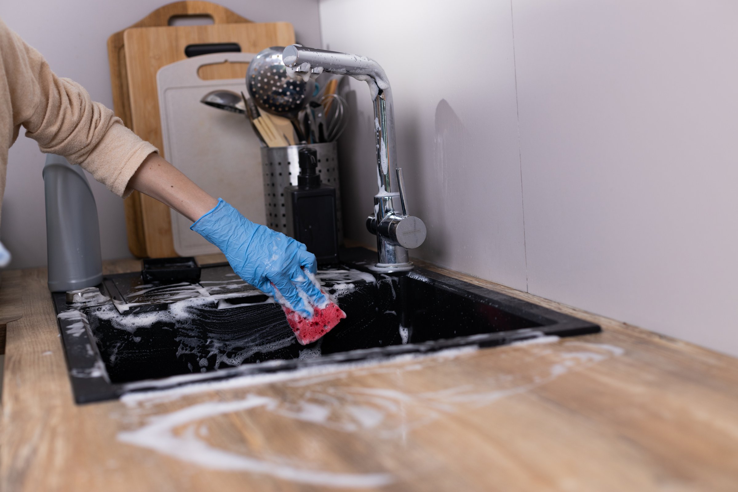 This image displays a person with a gloved hand using a sponge to clean a black kitchen sink. Bubbles and foam are visible, showcasing the cleaning process in a tidy kitchen.