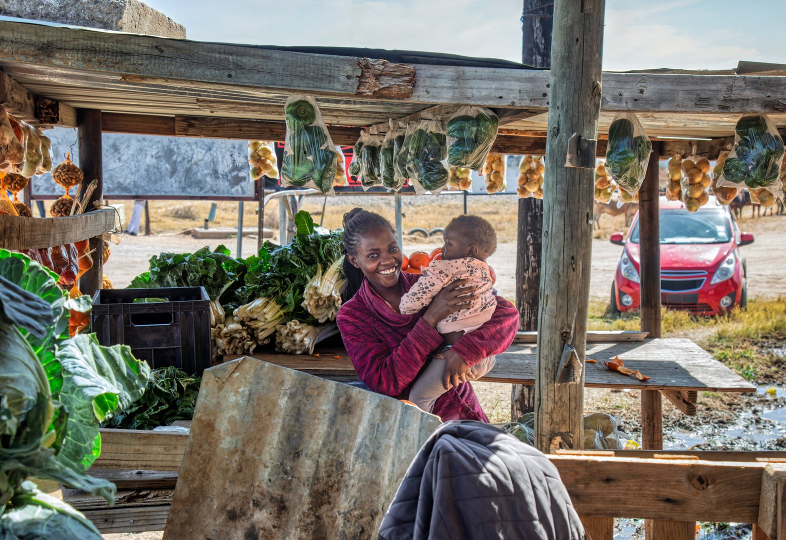 Happy African street vendor holding her child, shack with vegetables and fruits, women empowerment with skills Private Sector Engagement ,direct from farm sale