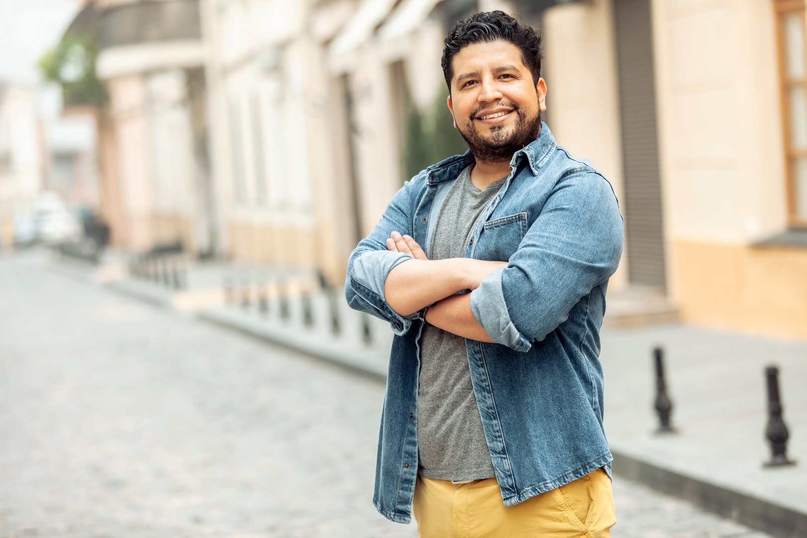 Smiling man in a denim jacket stands with arms crossed on a cobblestone street in an urban setting.