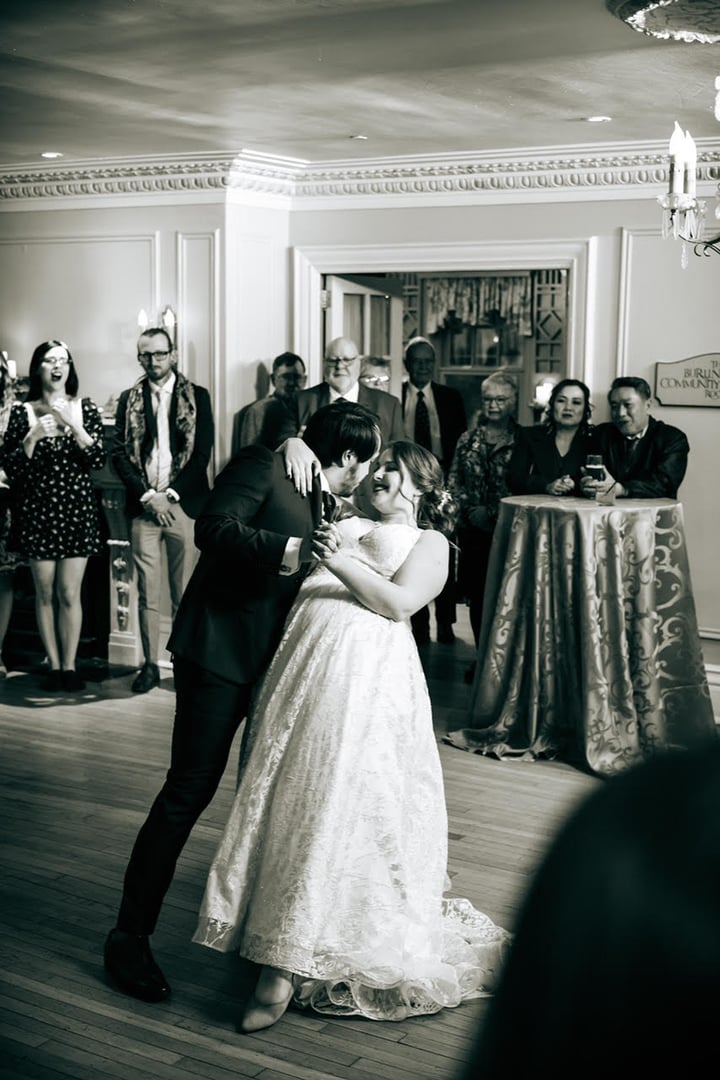 A bride and groom share their first dance, surrounded by smiling guests in an elegant ballroom setting.