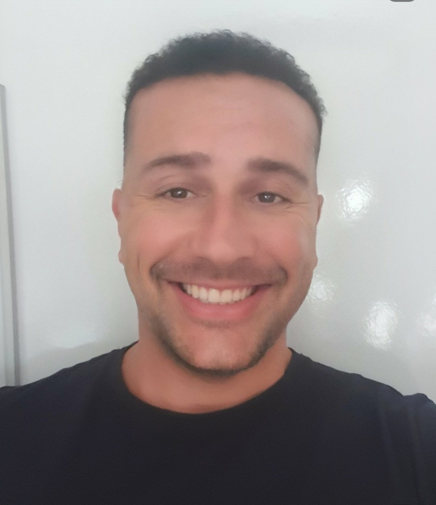 A man smiling broadly, wearing a black shirt, in front of a plain white background.