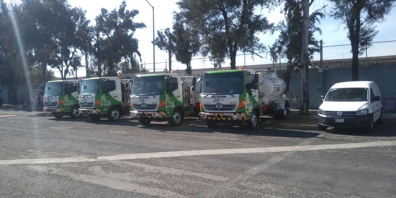 Row of green and white gas delivery trucks parked in a lot with trees and a fence in the background.