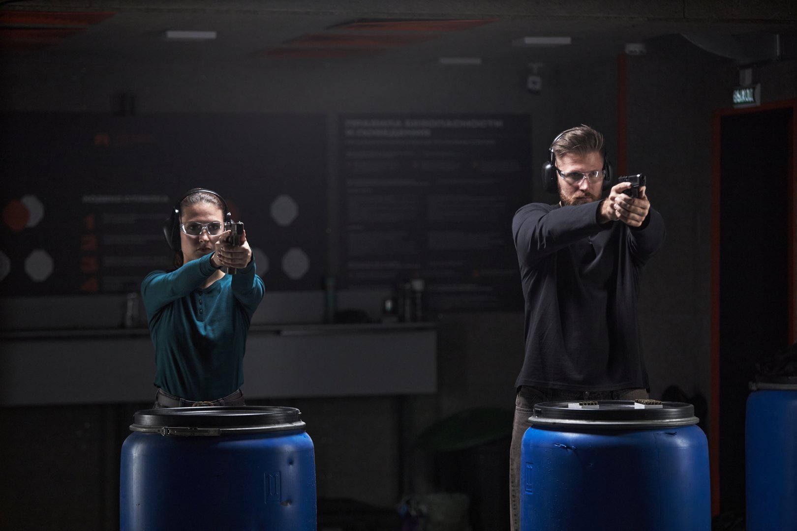 Caucasian young woman and Caucasian young man standing side by side aiming handguns at shooting range, both wearing protective glasses and ear protection, focusing on targets