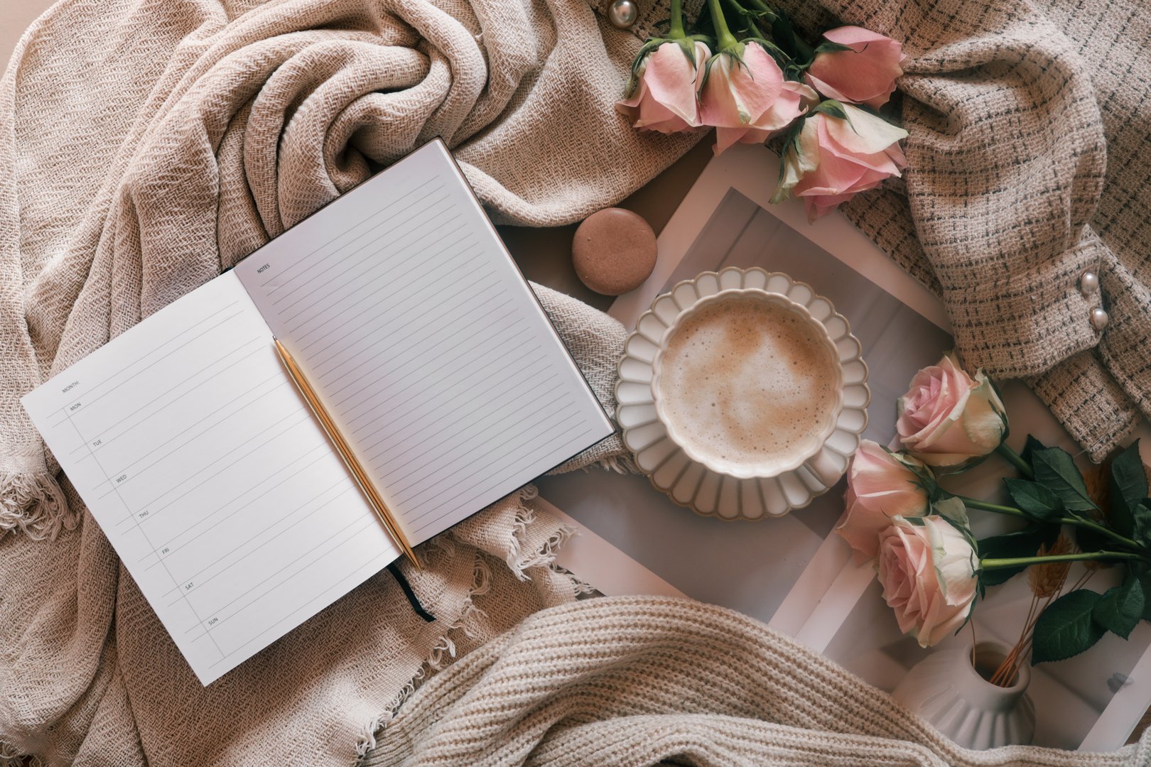 Flat lay of an open weekly planner with a pen, coffee cup, pink roses, and cozy neutral fabrics. Ideal for planning, journaling, or productivity lifestyle themes with a soft feminine aesthetic.