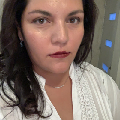 A woman with dark hair wearing a white blouse with lace details, looking directly at the camera.