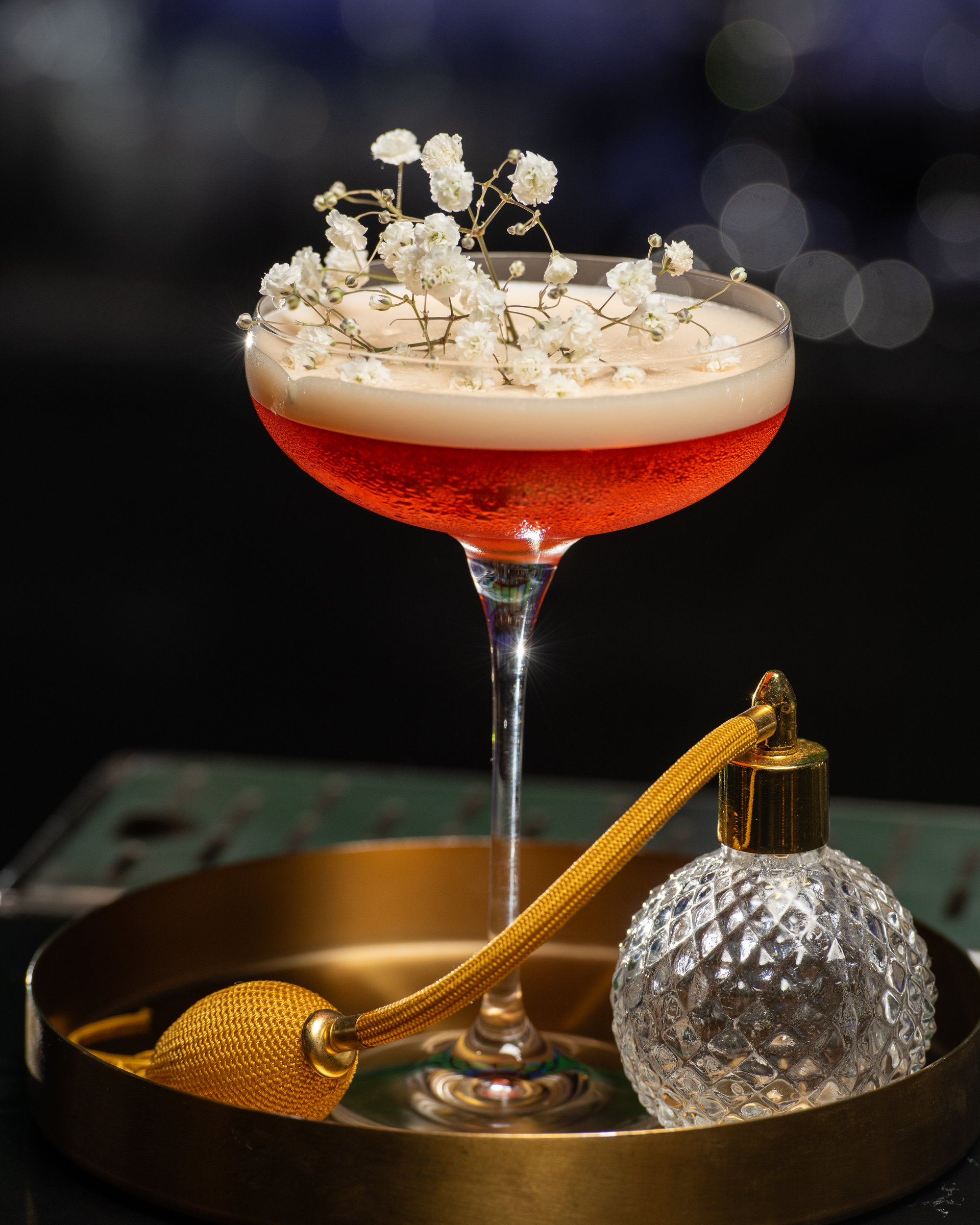 blush-toned cocktail with a frothy top is elegantly garnished with delicate white baby's breath flowers, presented on a golden tray alongside a vintage-style perfume atomizer.