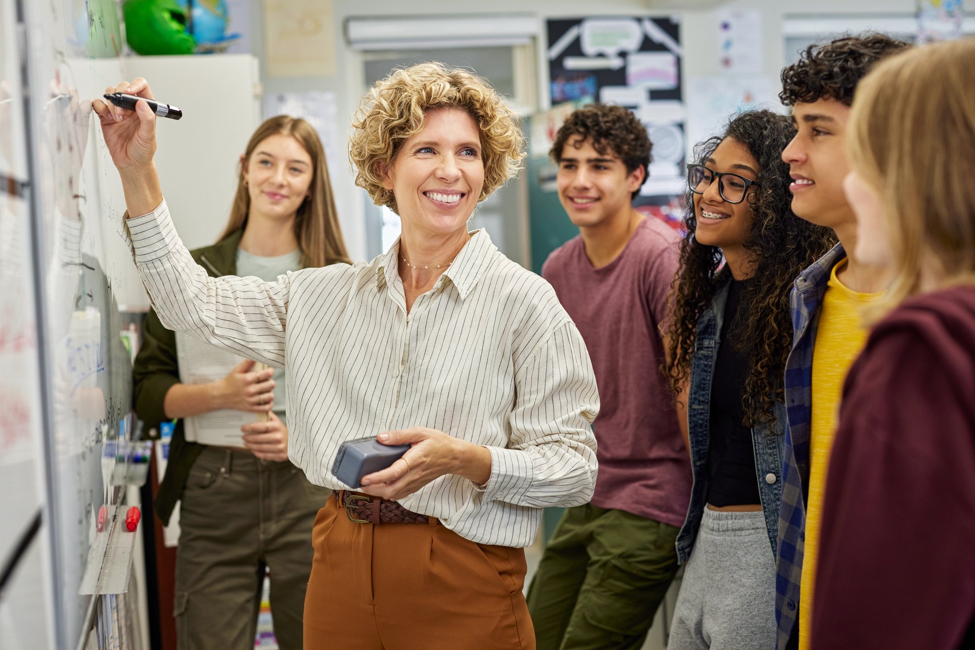 Smiling happy teacher writing on whiteboard during lesson at secondary school. Group of multiethnic students watching and smiling while learning near professor. Interactive classroom during learning activity at high school.