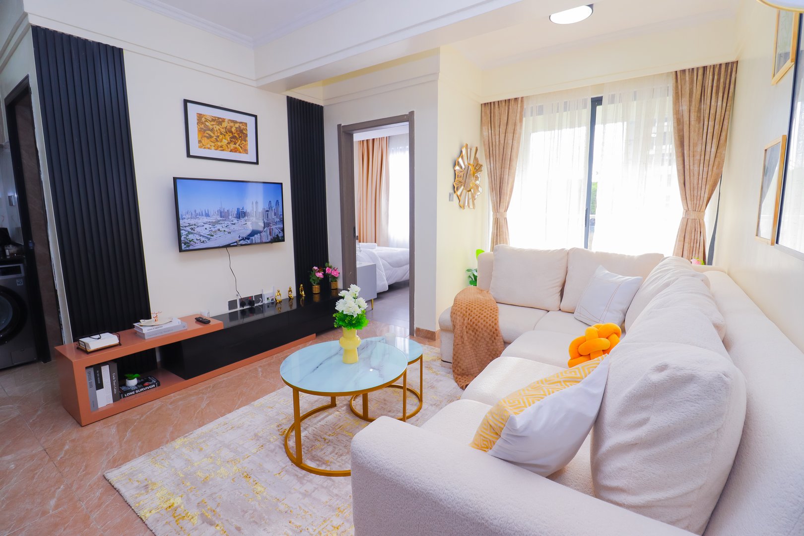 Modern living room with light sofa, round glass coffee table, TV, and art on walls, featuring natural lighting from windows.