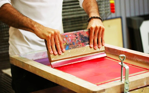 Person using a squeegee to apply red ink on a screen printing frame.
