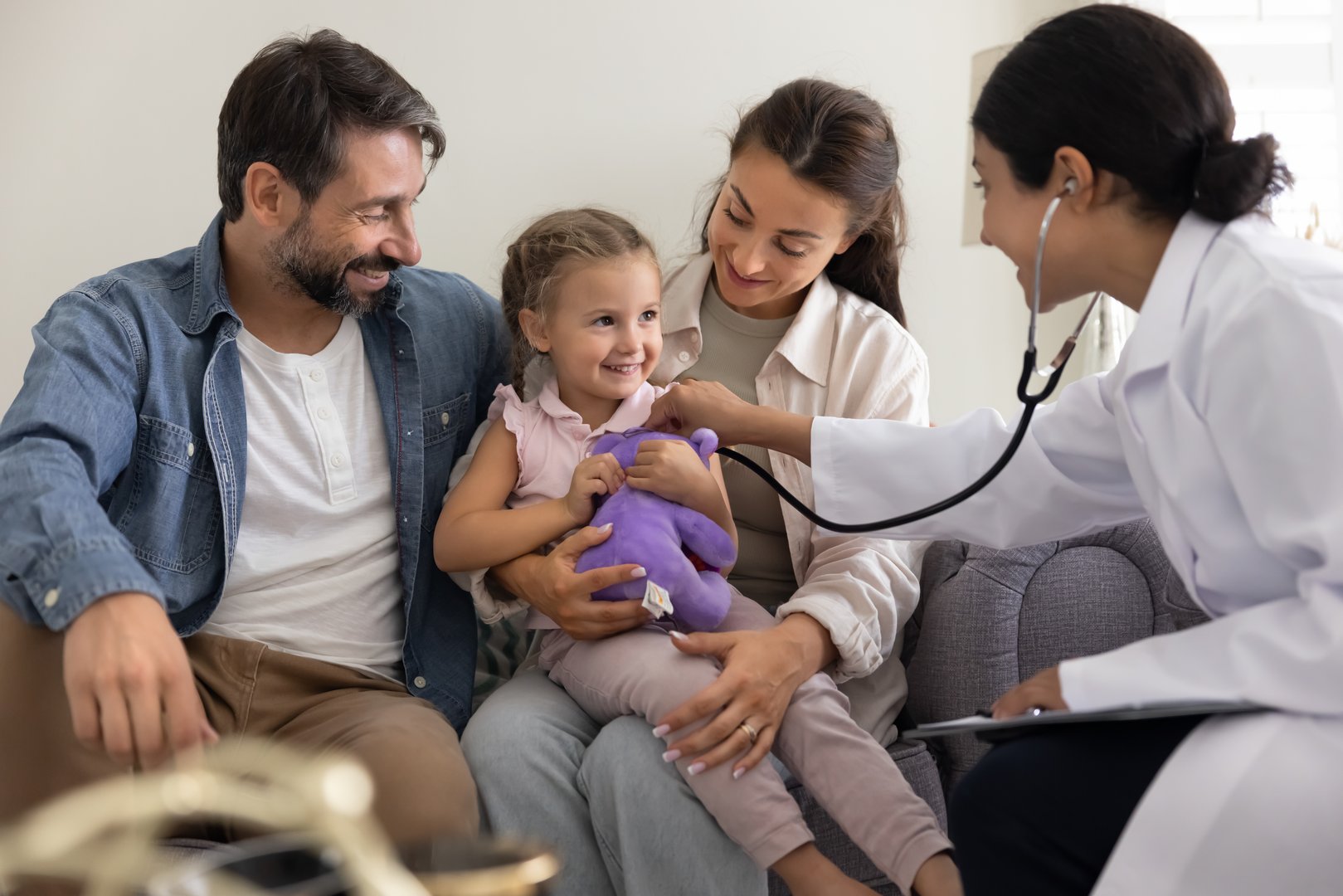 Cute 3s girl sitting on mum lap with favourite fluffy toy, while Indian female doctor in white coat listening her heartbeat using stethoscope. Regular check to prevent illness, healthcare, pediatrics