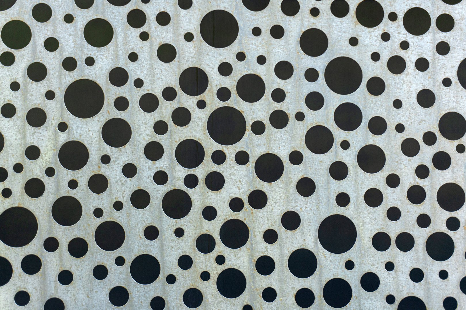 Galvanized metal sheet with randomly spaced round black holes of varying diameters. Light traces of rust are visible on the surface. Background.