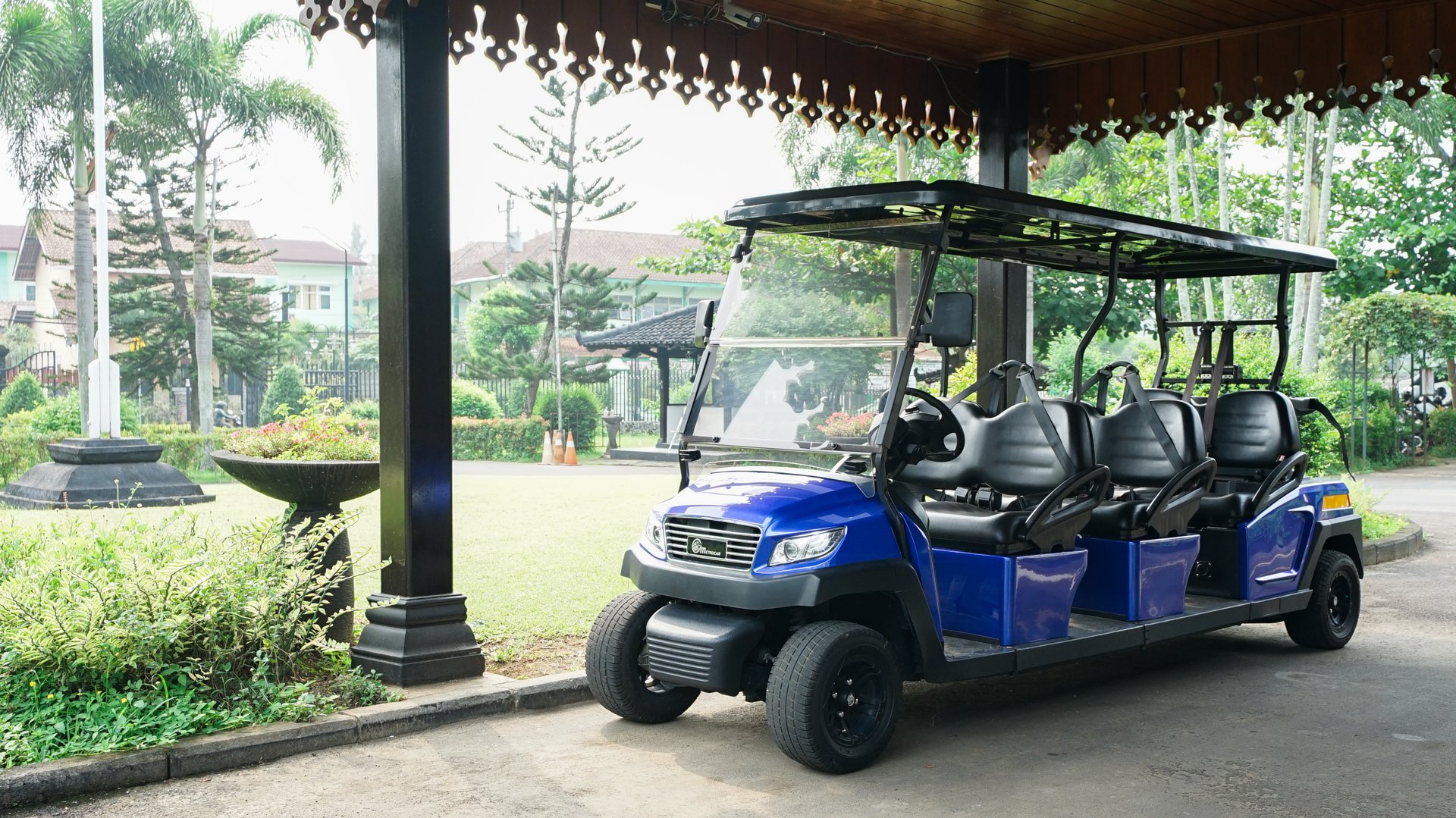 A modern electric golf cart with a sleek blue design is parked under a traditional pavilion. The open-sided structure features wooden carvings, providing a contrast between modern transportation and classic architecture. Lush greenery and urban surroundings complete the serene outdoor setting