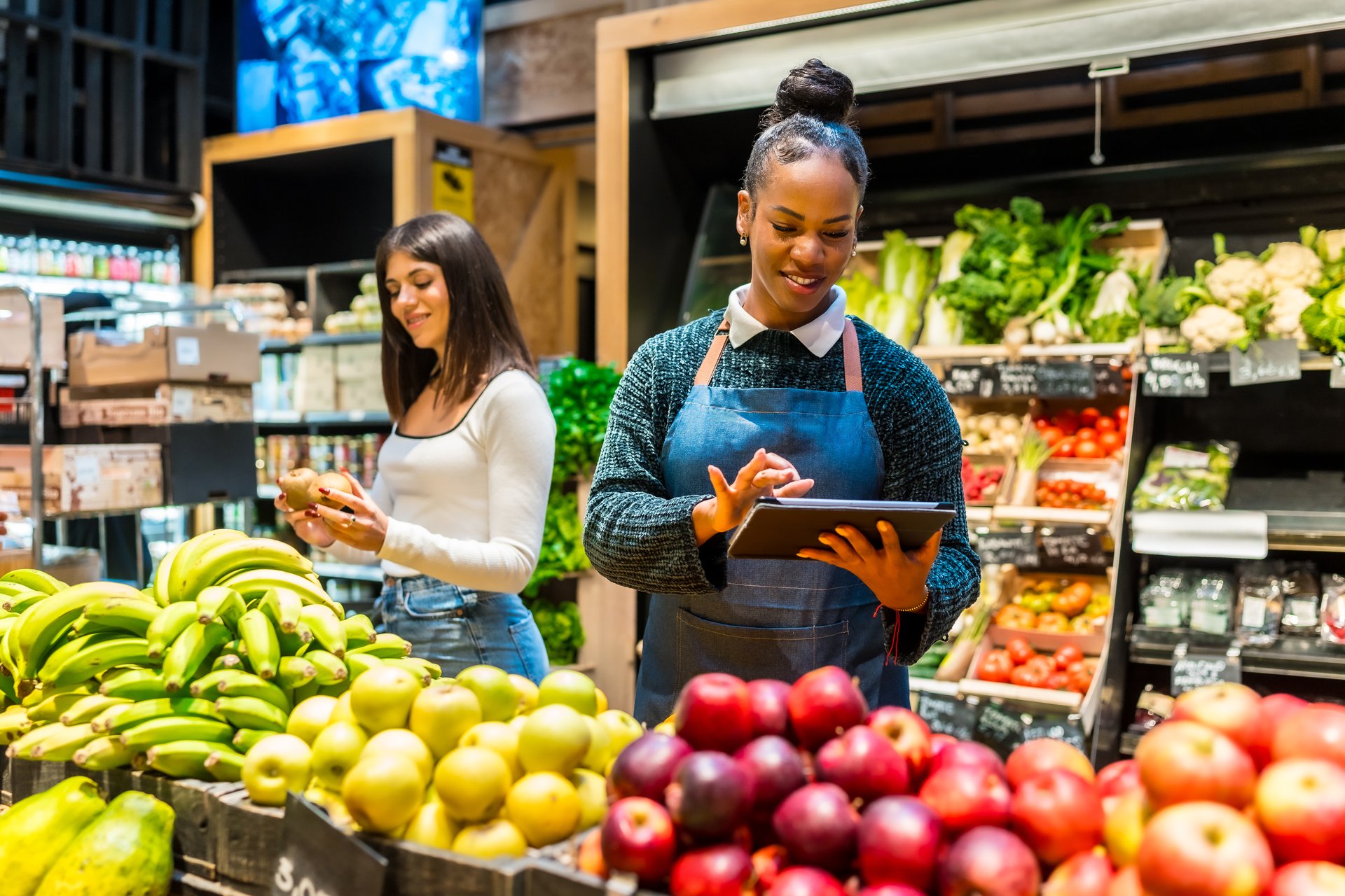 Smiling saleswoman using digital tablet while working at organic supermarket, customer choosing kiwi fruit