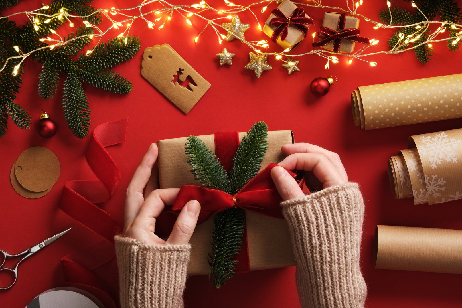Women's hands tying ribbon on a gift. Table with handmade supplies, rolls of kraft brown paper, red velvet ribbon, labels, scissors.