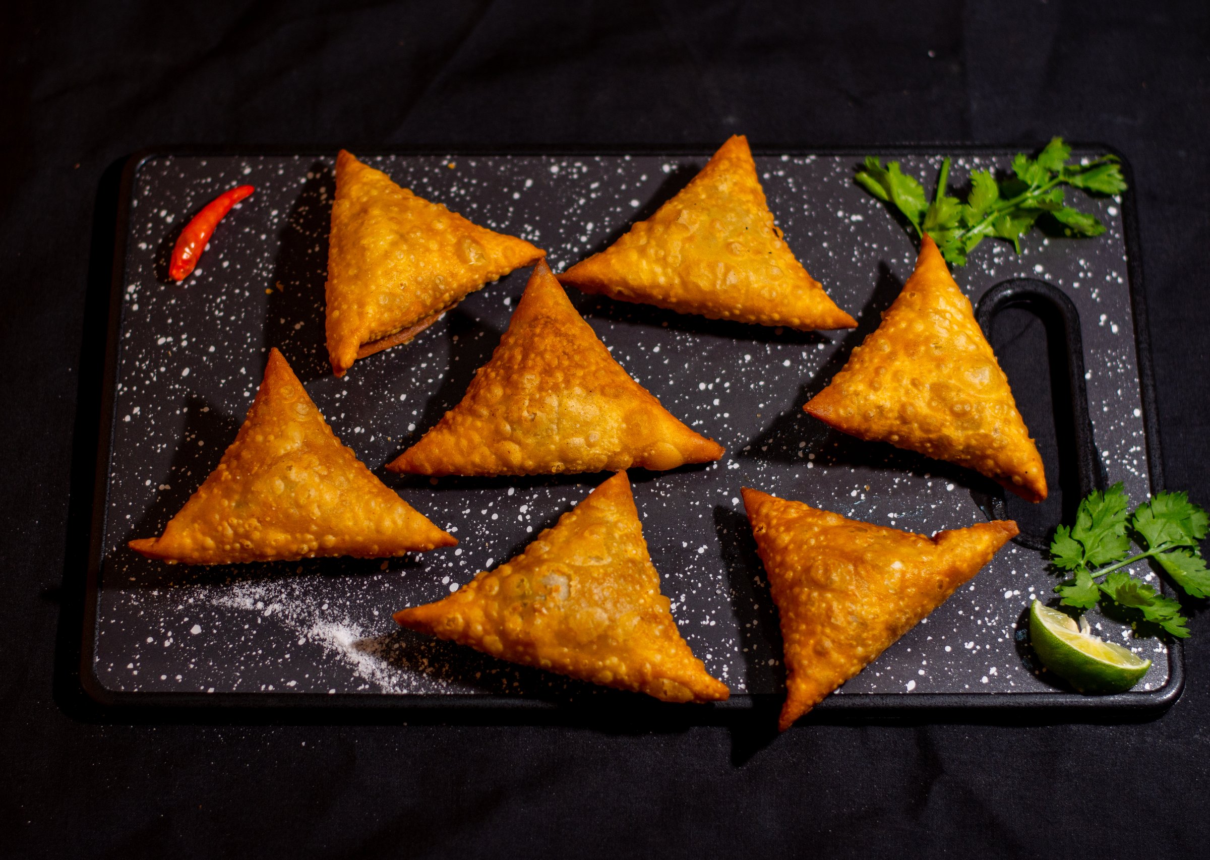 Seven fried samosas arranged on a black cutting board with chili, cilantro, lime wedge, and sprinkled flour.