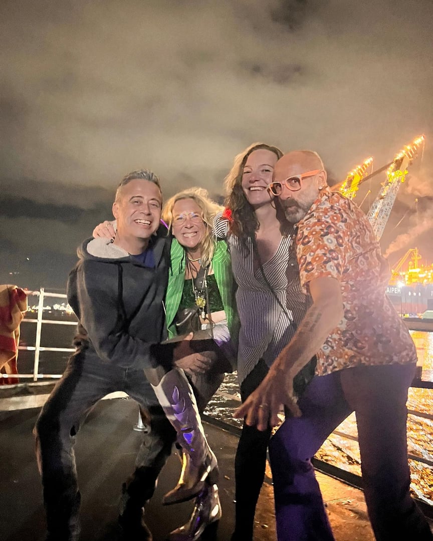 Four people posing happily on a boat at night with city lights and cranes visible in the background.