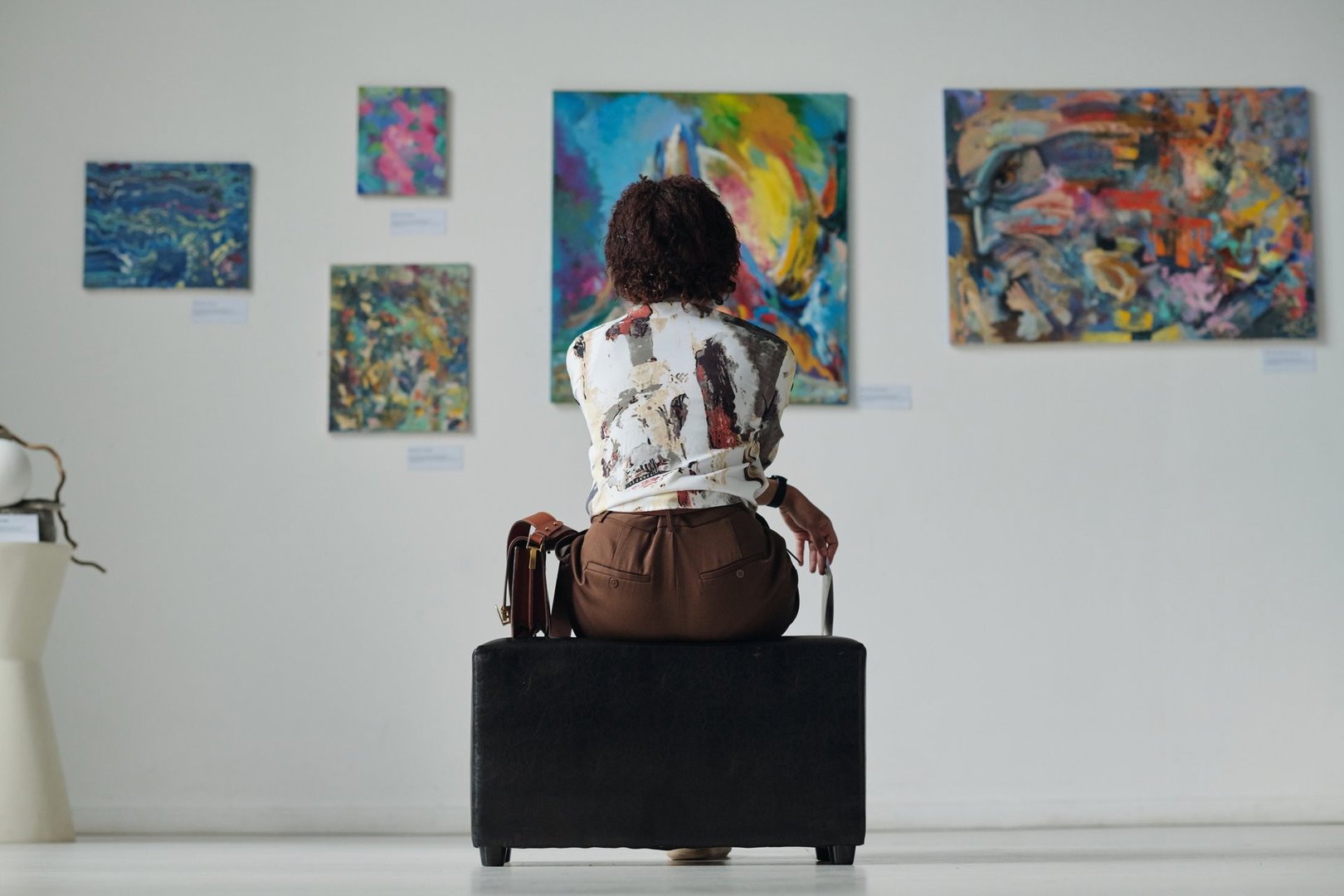 Rear view of young woman sitting on couch and looking at paintings on the wall, she enjoying art in gallery