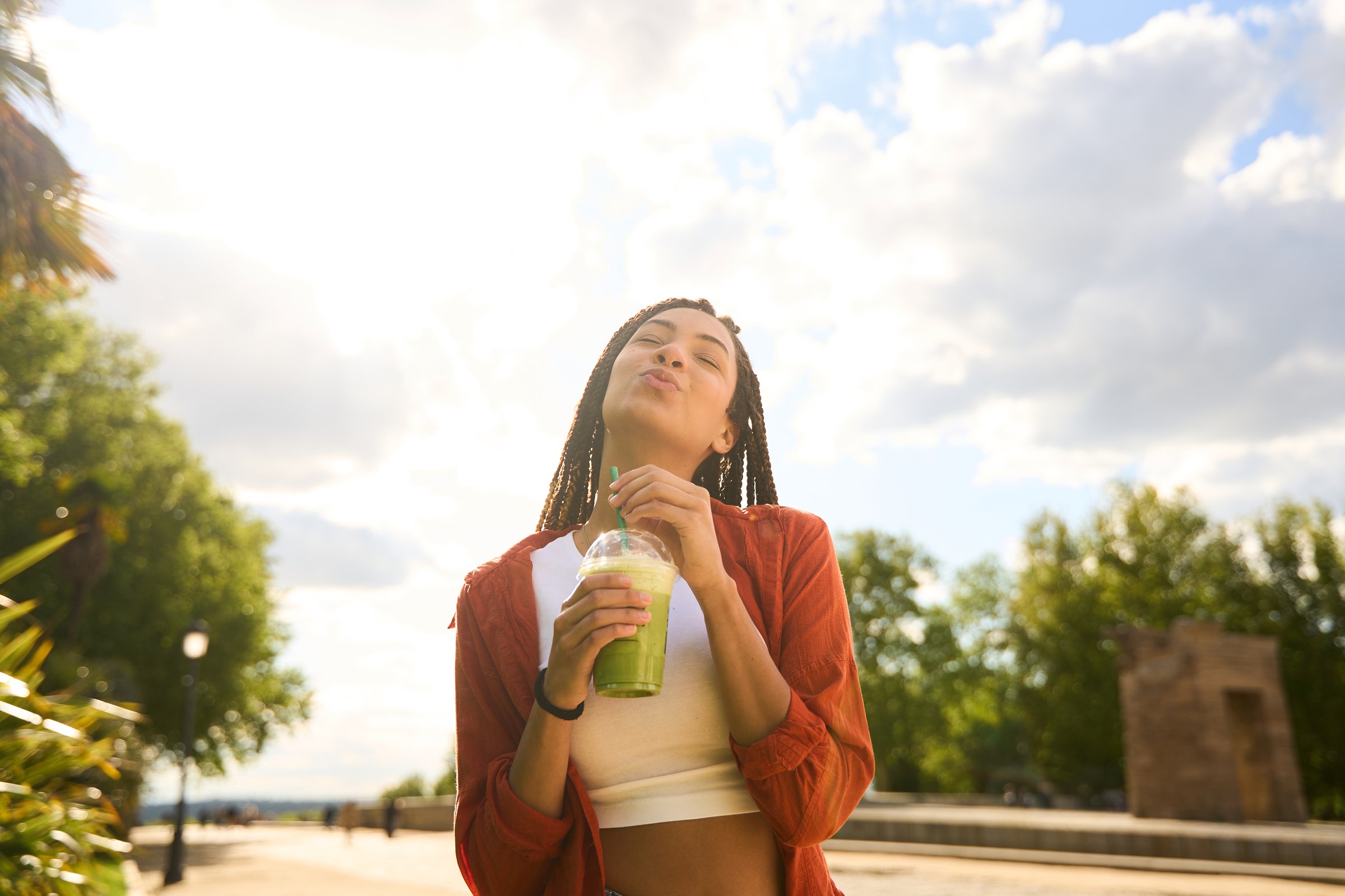 Young woman with braids savoring a refreshing green smoothie while basking in the sun on a beautiful day at the park