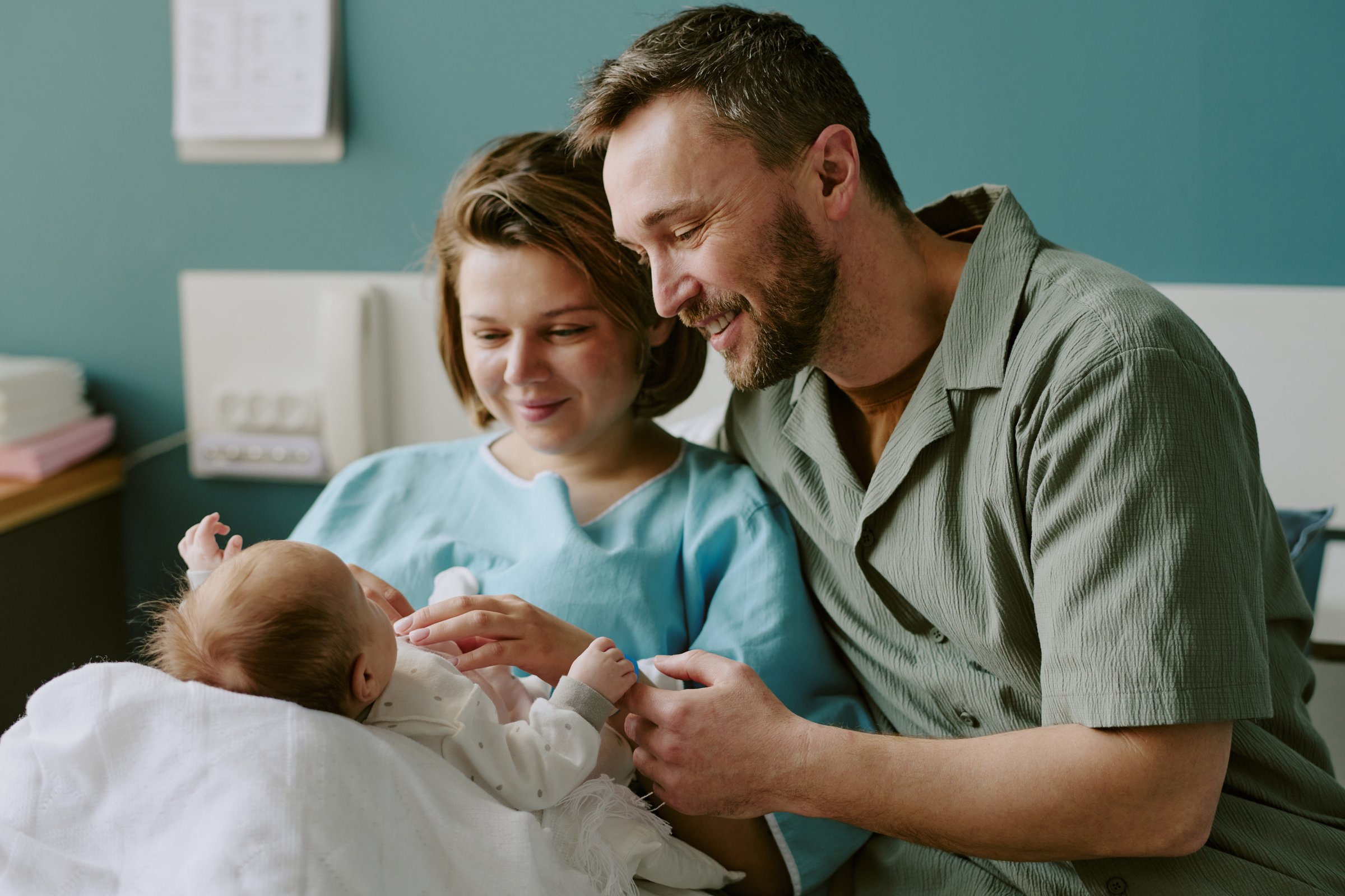 Smiling parents holding and admiring their newborn baby in hospital room. New parents gazing lovingly at infant wrapped in white blanket and enjoying moment together