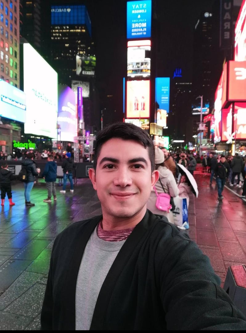Person taking a selfie in Times Square at night with bright billboards and a crowd in the background.